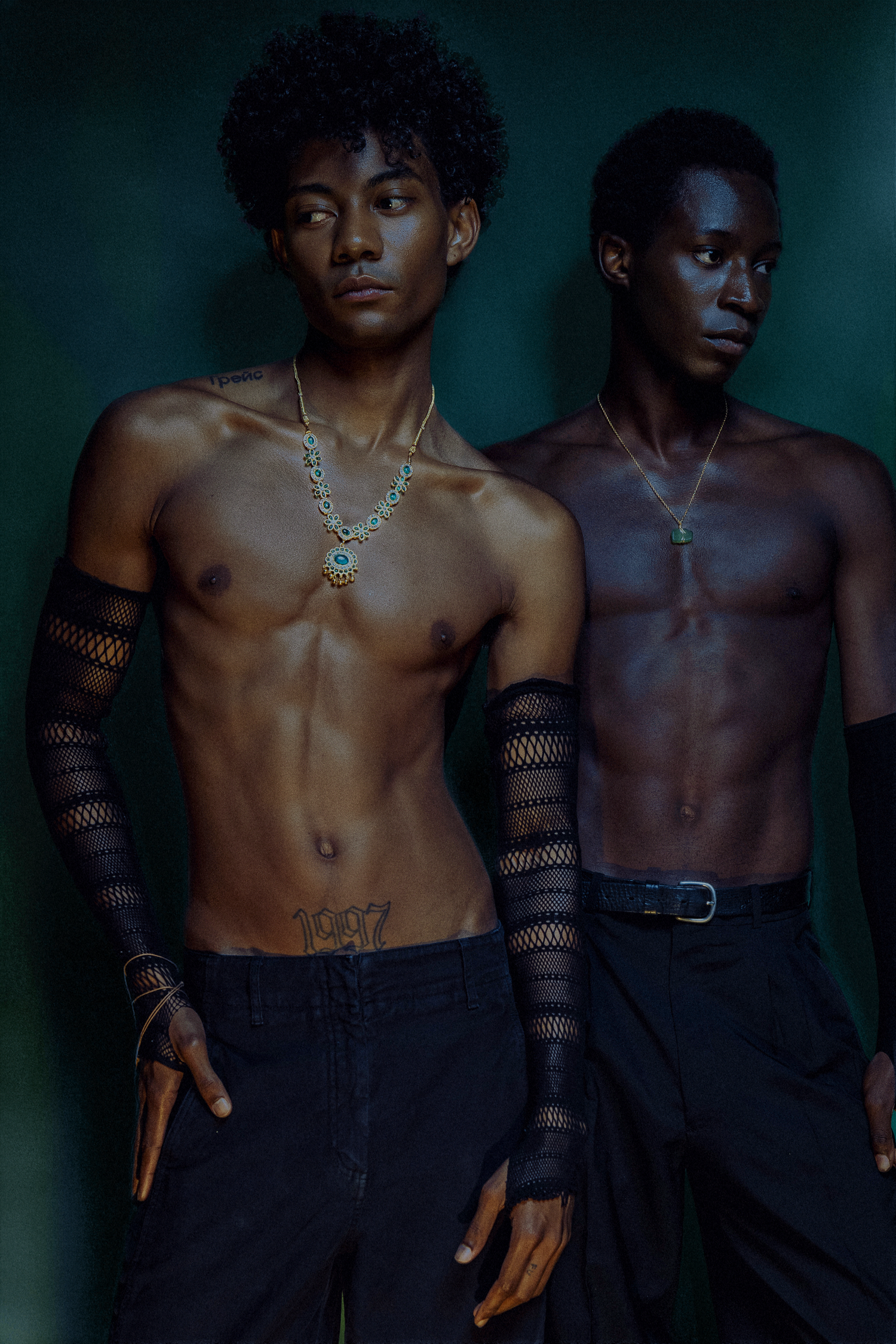 Two shirtless Black male models wearing lace arm sleeves and necklaces pose against dark green backdrop.