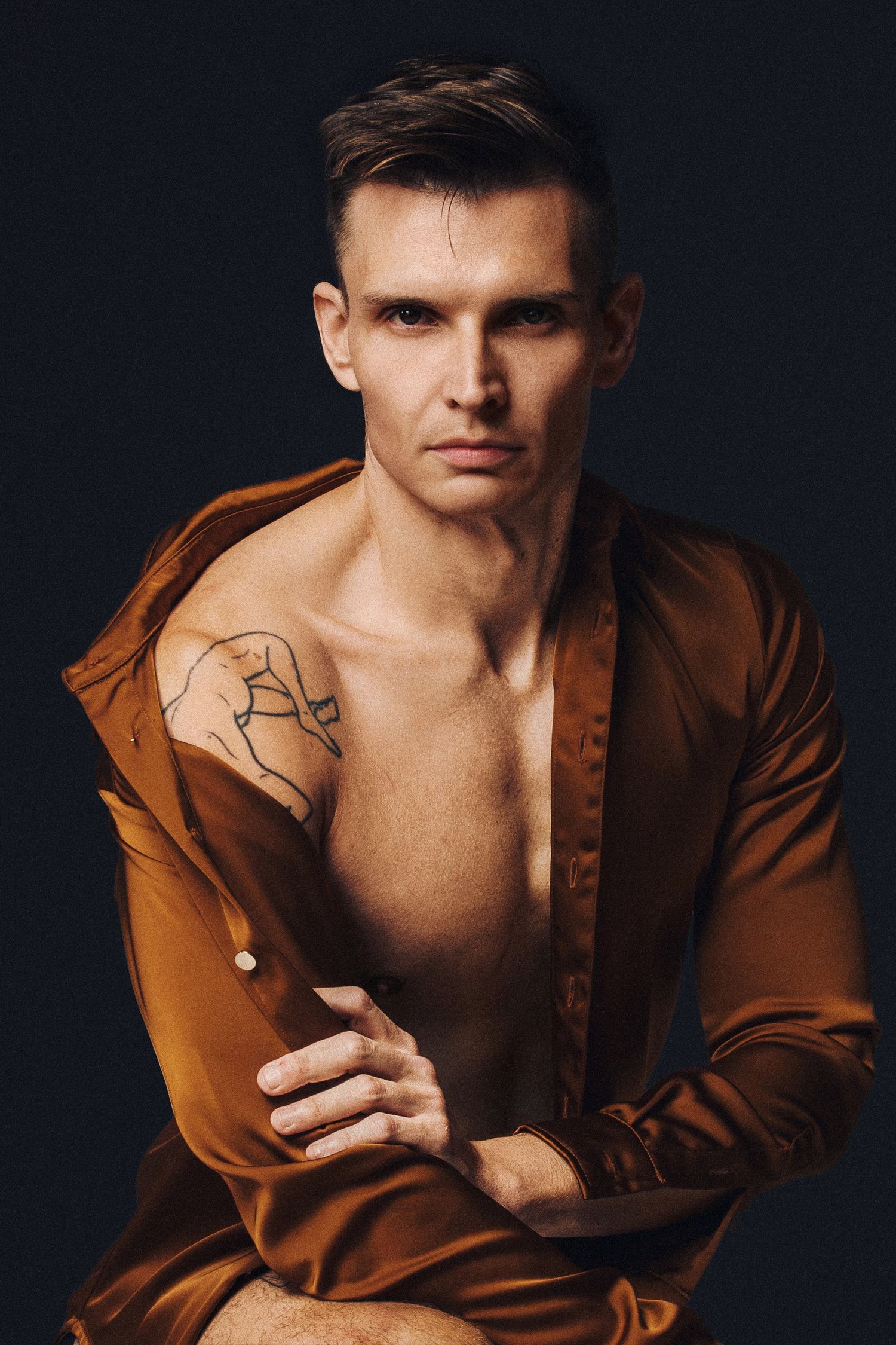 Muscular man in open bronze satin shirt with shoulder tattoo poses confidently against dark background.