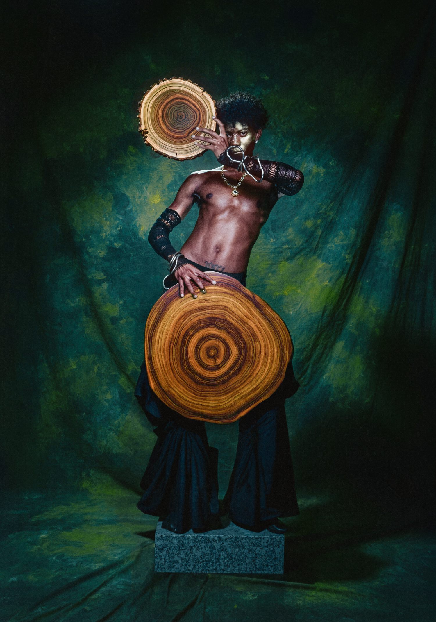 Shirtless model holds woven circular shields in dramatic pose against green backdrop.