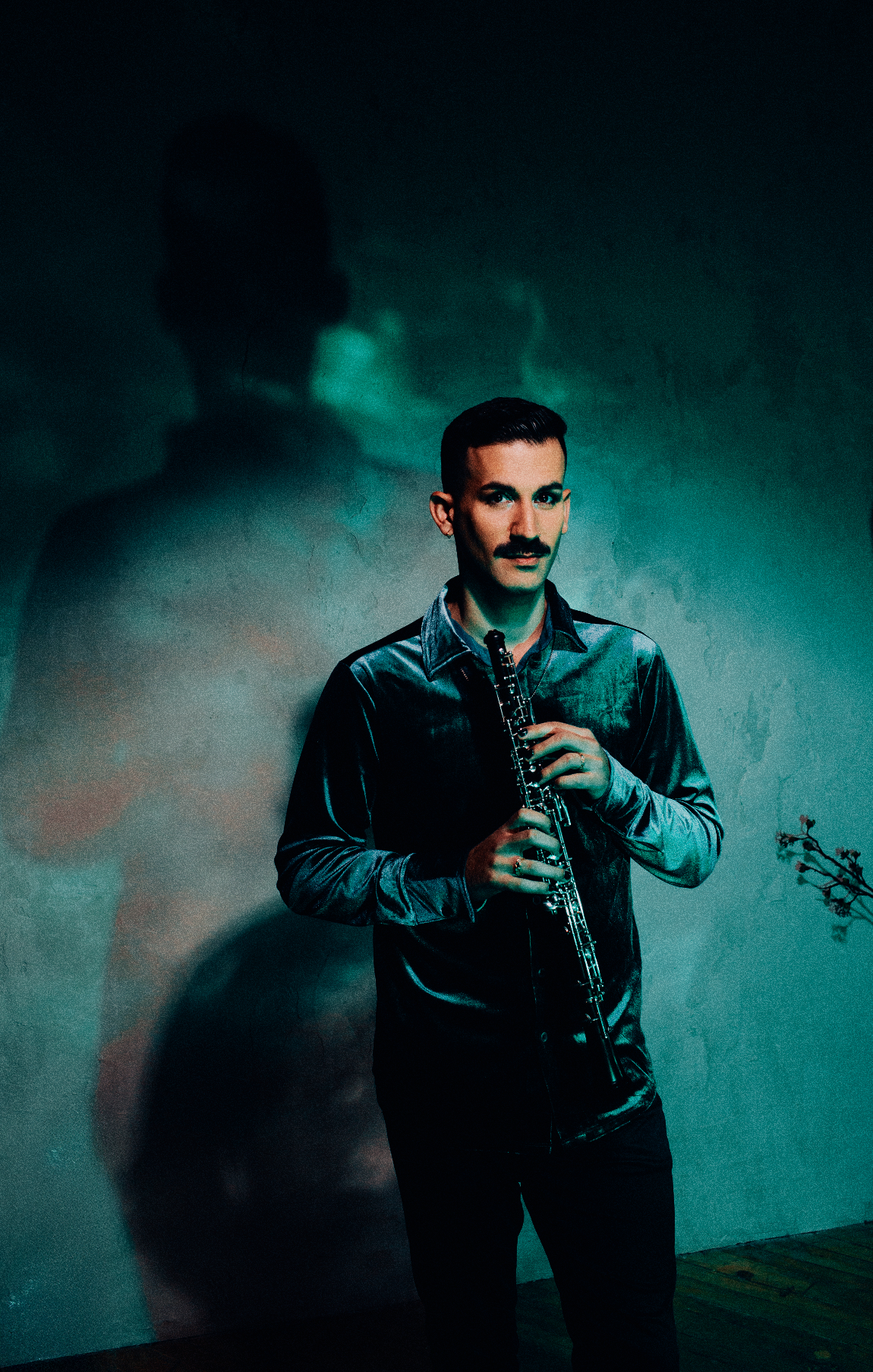 Musician in teal velvet shirt holds clarinet against moody teal-lit wall with dramatic shadow.