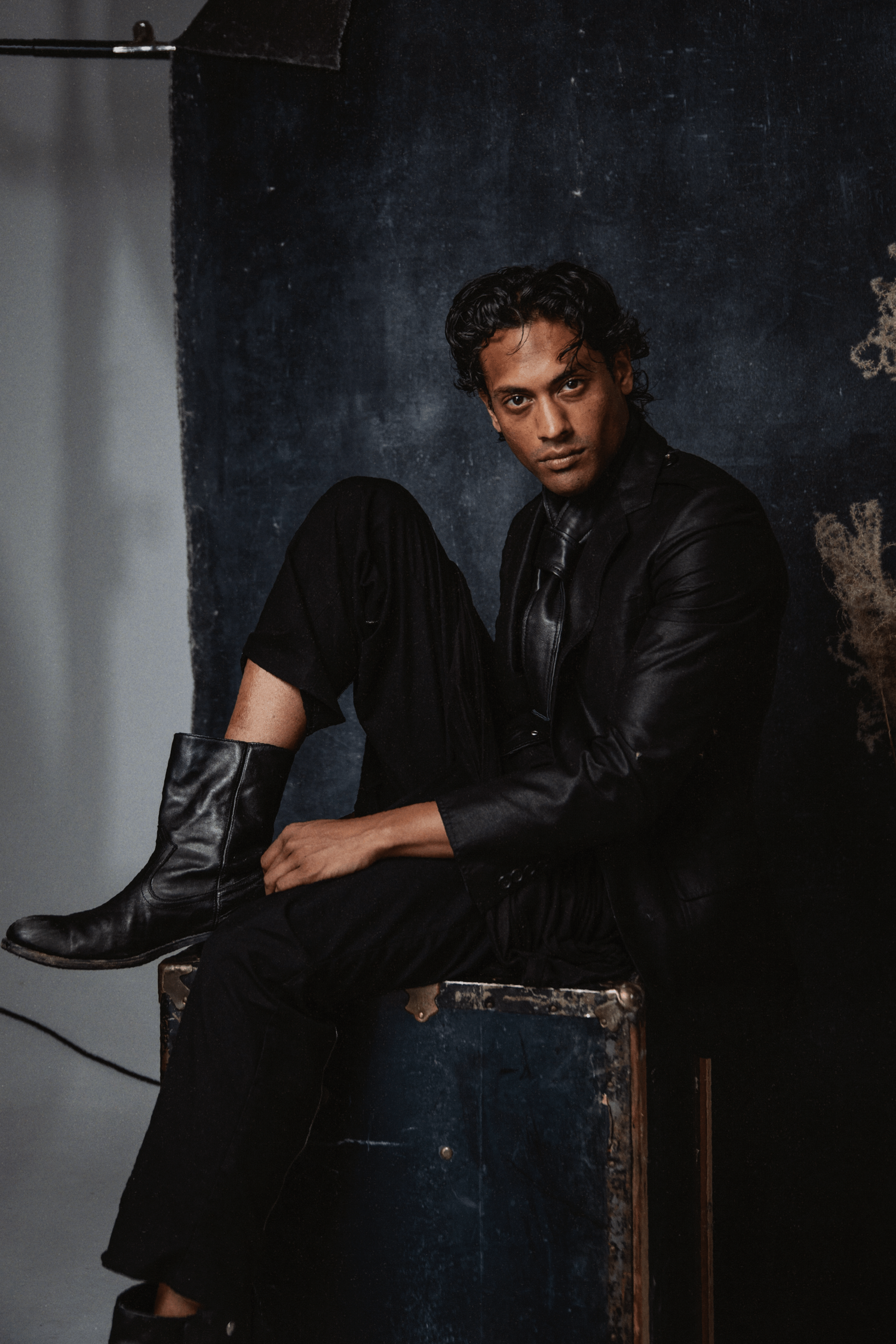 Young man in all-black leather jacket and boots seated on vintage trunk against dark textured backdrop.