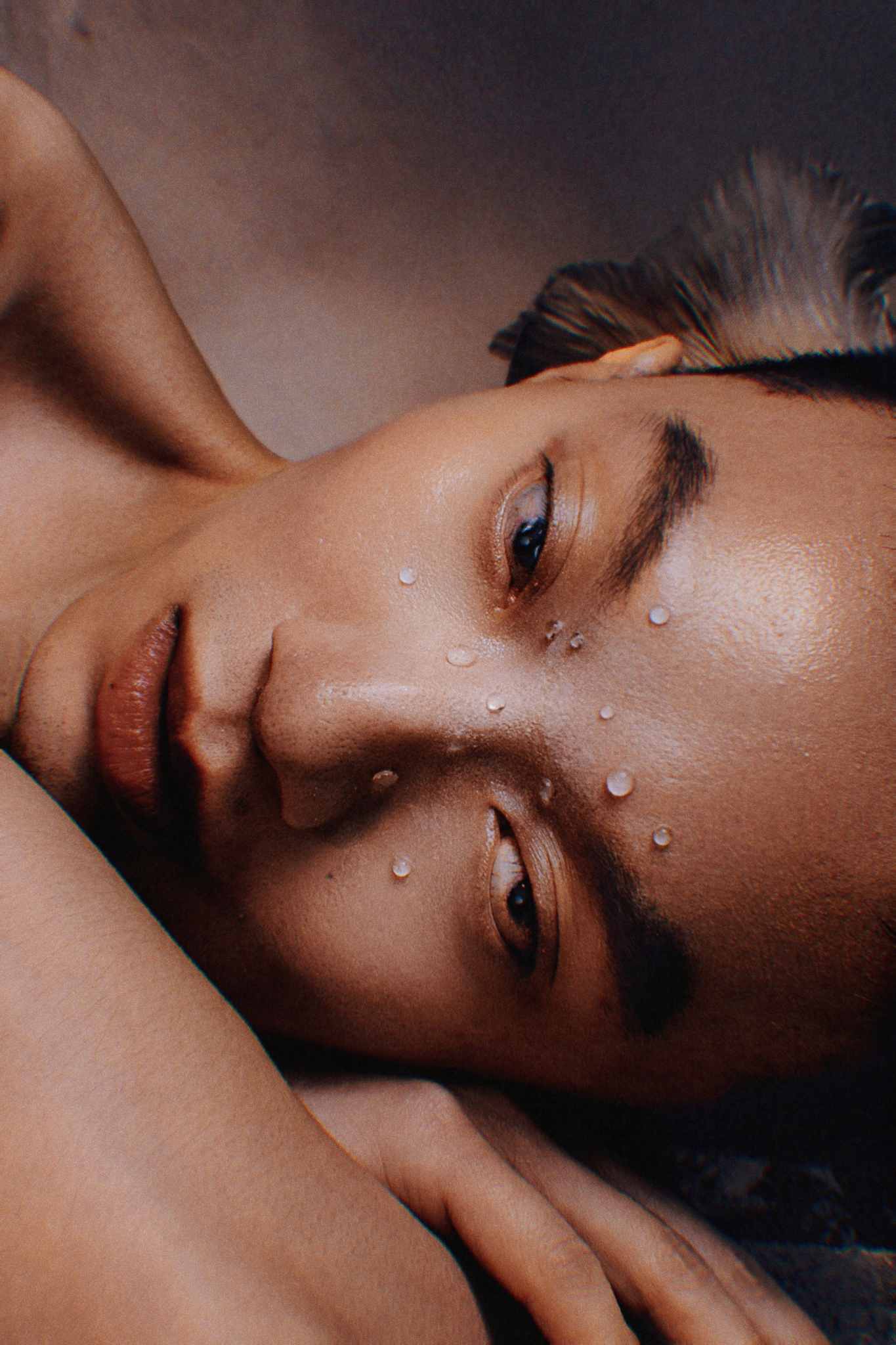 Close-up portrait of a person lying down with water droplets on glowing skin in moody lighting.