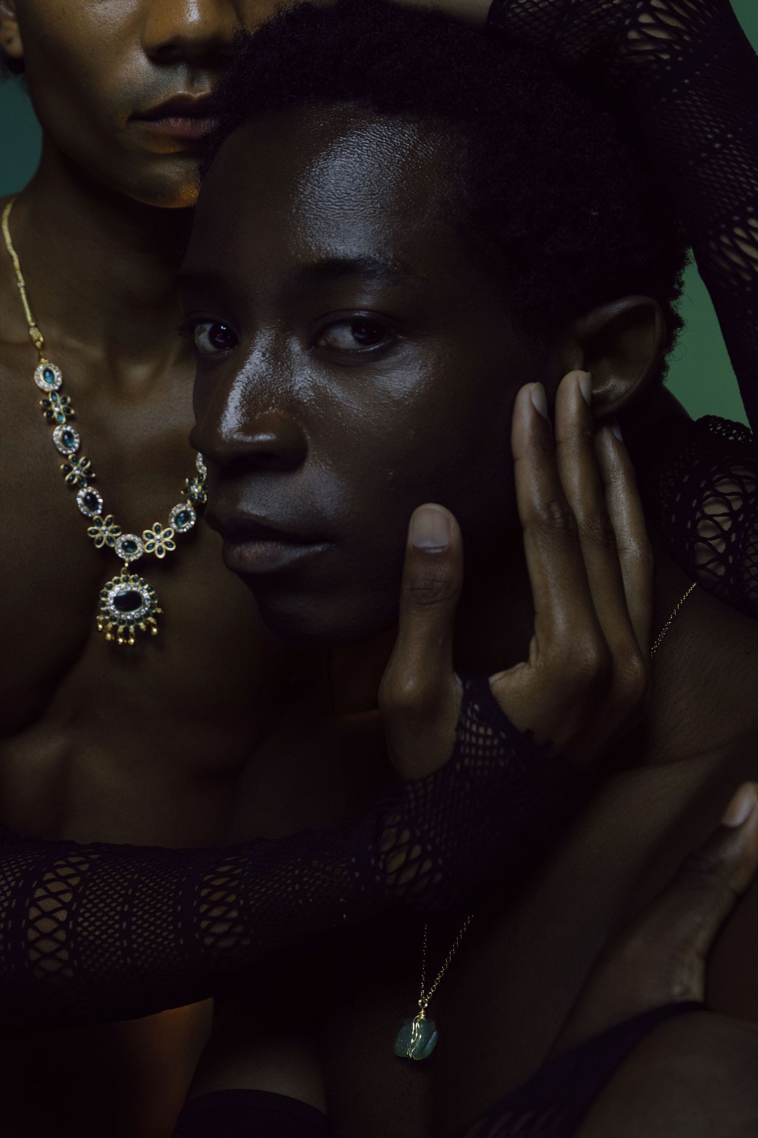 Two Black models wearing elegant gold gemstone jewelry posed together against a dark green background.