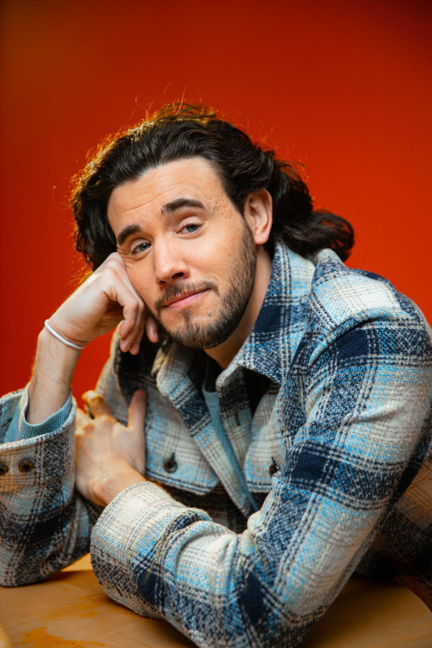 Bearded man with dark hair in plaid jacket resting chin on hand against vibrant red background.