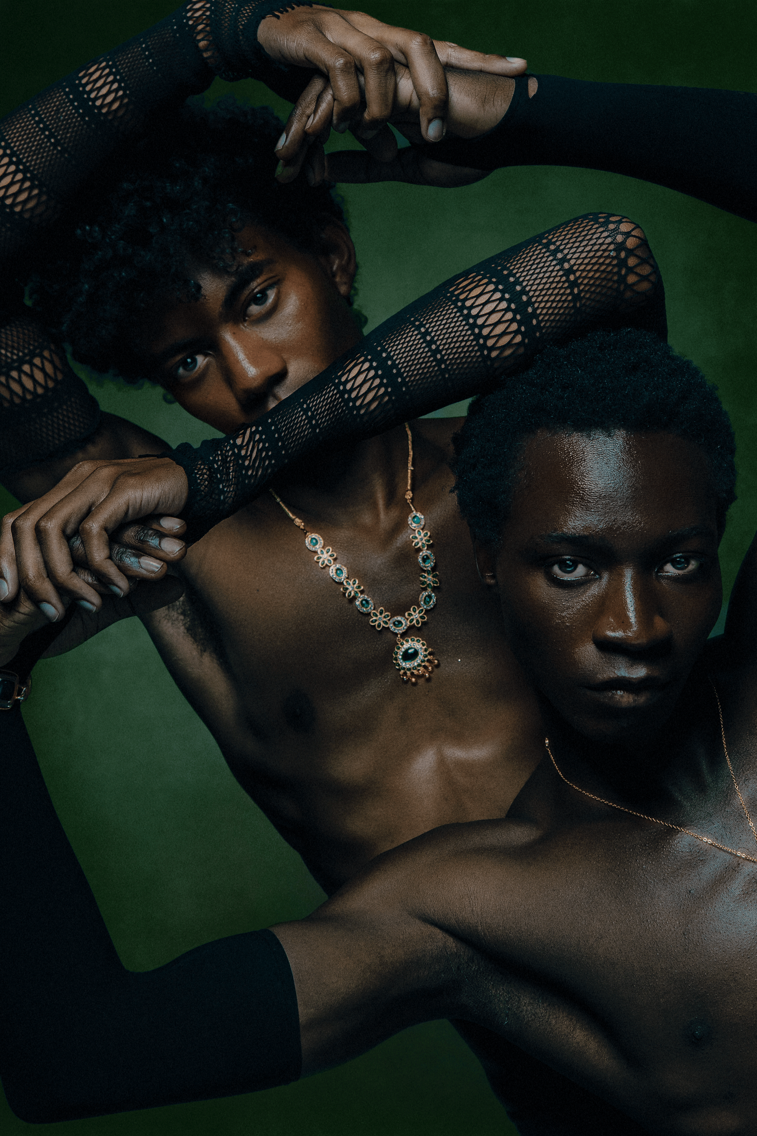 Two Black male models in striking pose against dark green background, styled with mesh sleeves and jewelry.