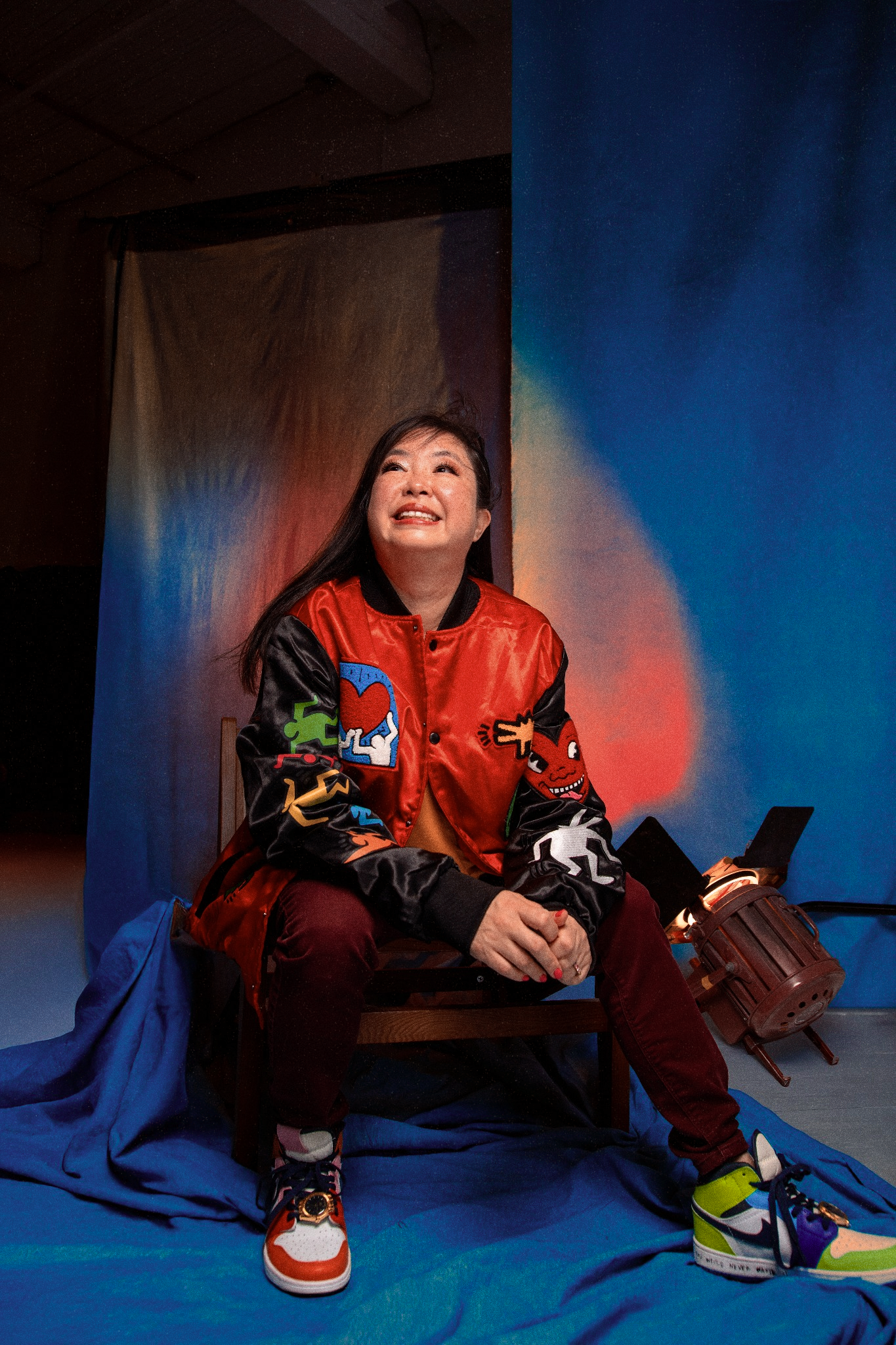 Woman in red bomber jacket seated on chair against a colorful draped backdrop.