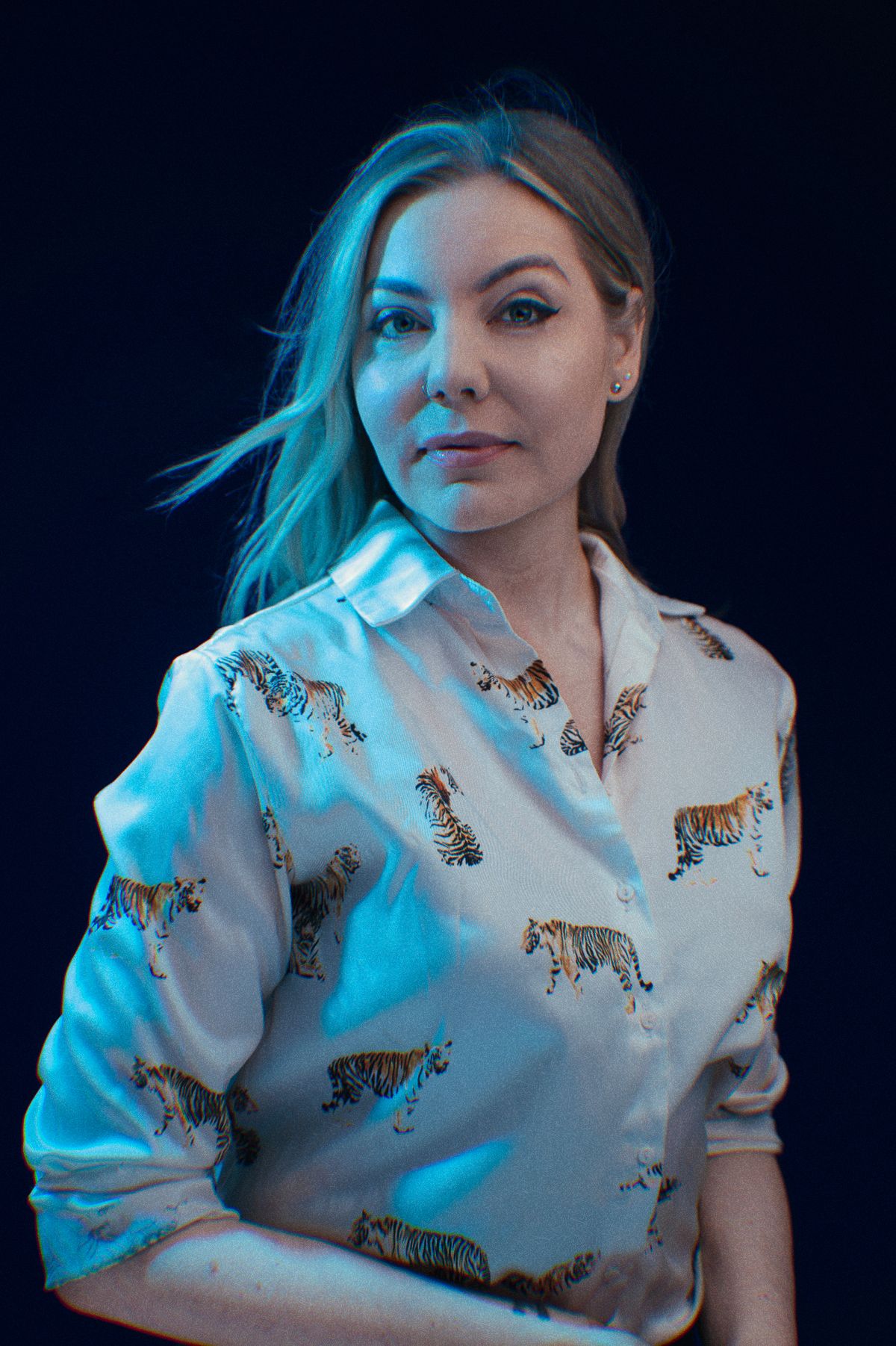 Stylish blonde woman wearing a white tiger-print button-up shirt poses in blue-toned studio lighting.