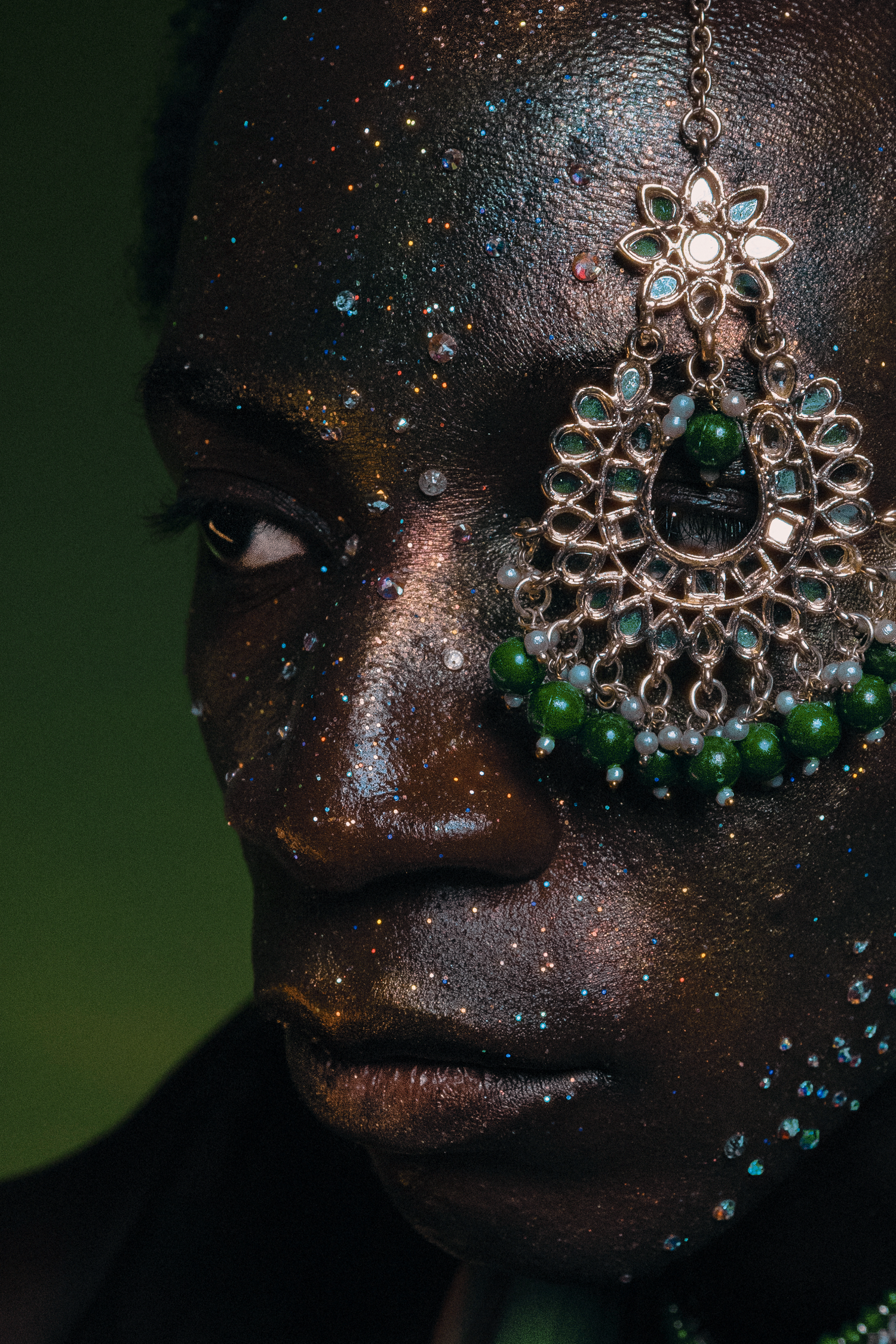 Close-up of a Black model with glitter makeup wearing an ornate green and gold chandelier earring.