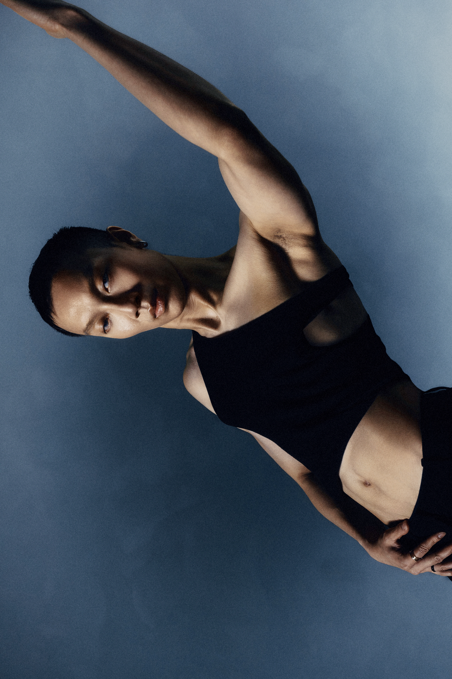 Athletic model in black crop top stretches arm overhead in dynamic pose on blue background.