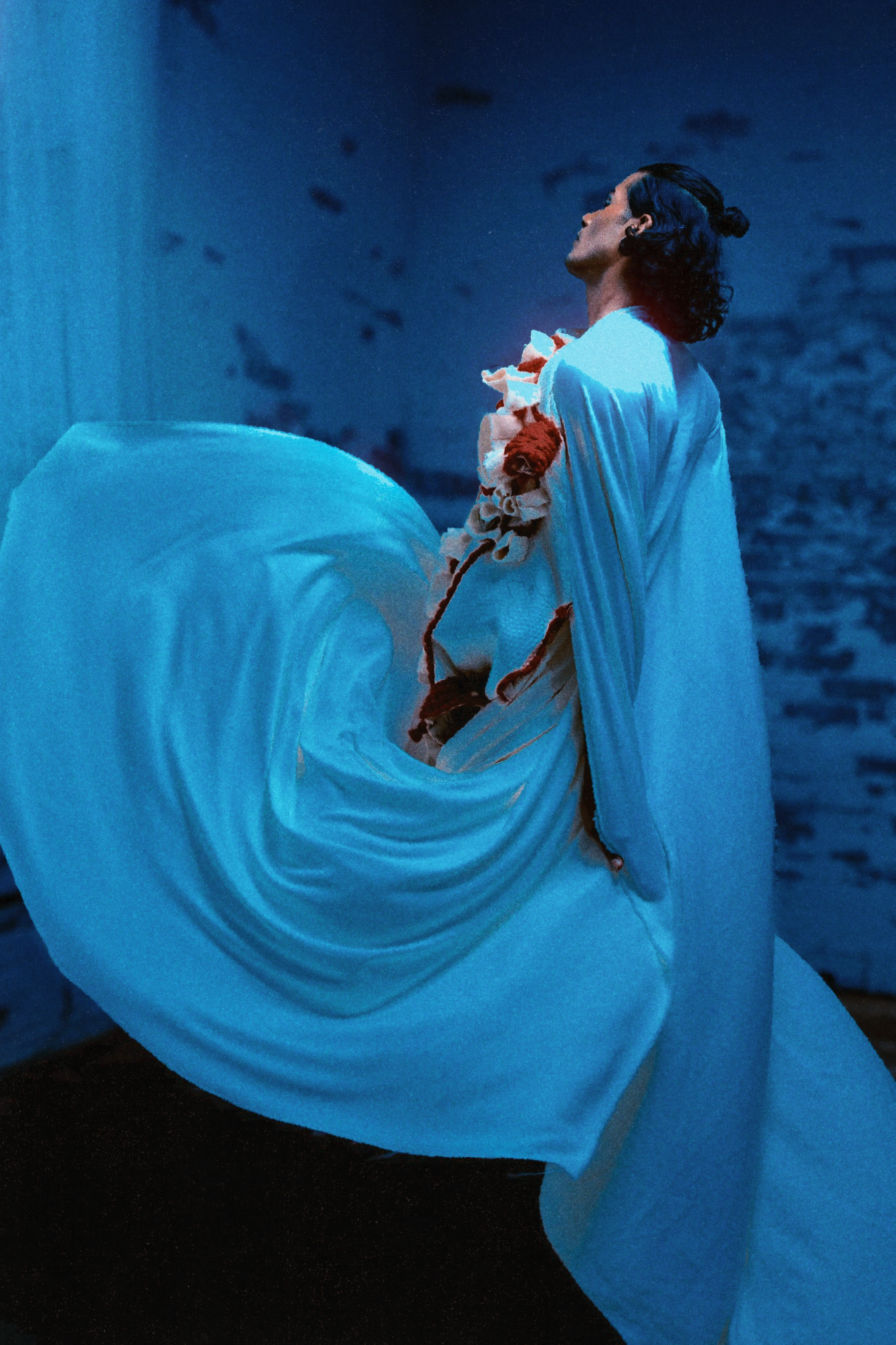 Figure draped in billowing cyan fabric holding cherry blossoms in moody blue-lit setting.