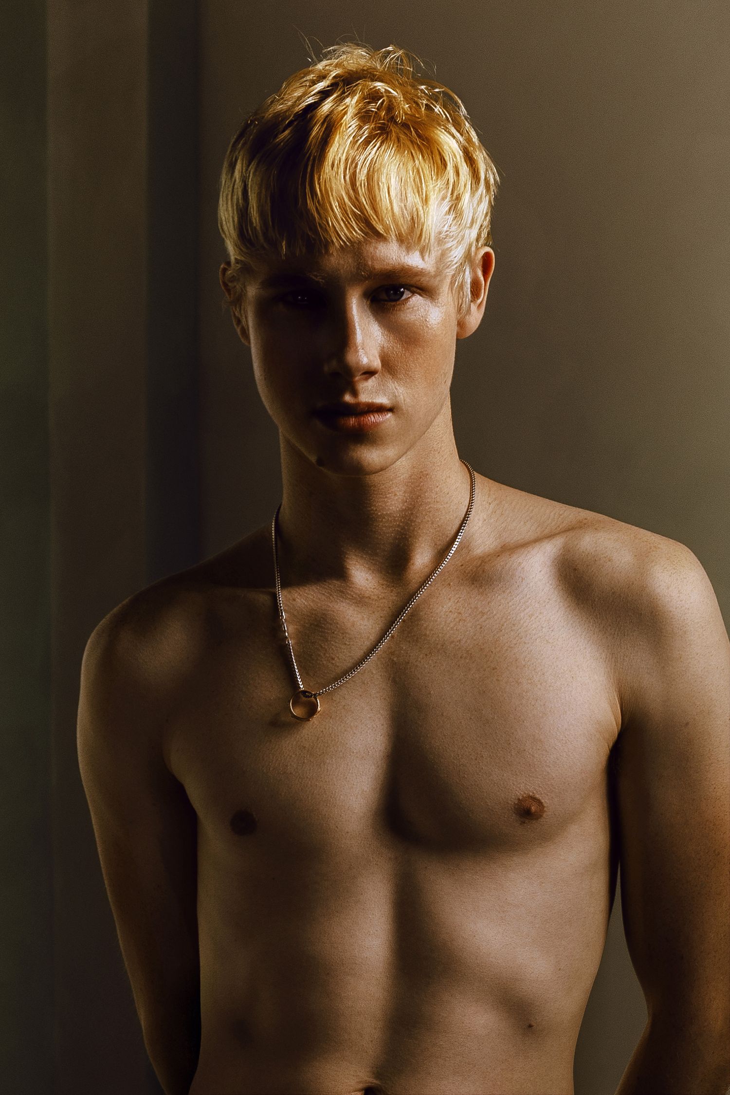 A shirtless young man with blonde hair wearing a silver chain necklace poses against a dark studio backdrop.
