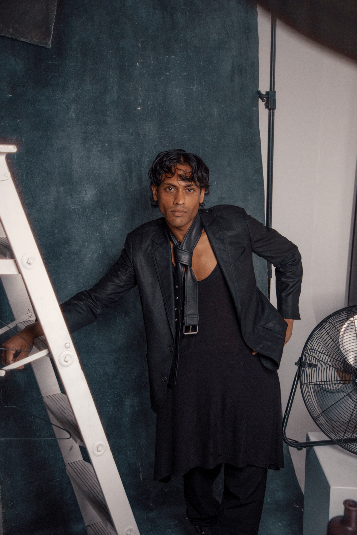 Stylish man in black outfit leaning on ladder against dark teal backdrop in photography studio.