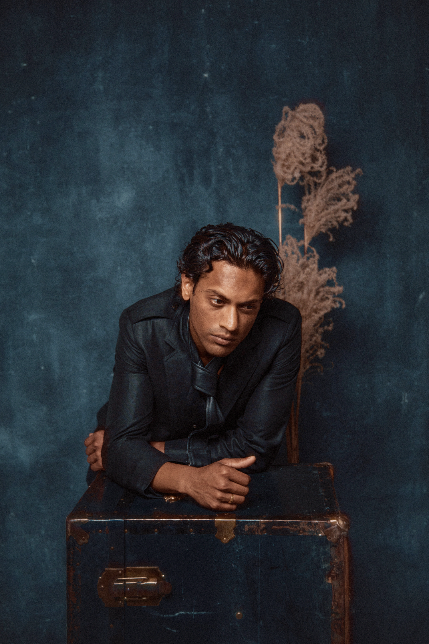 A South Asian man in a black leather jacket rests arms on a vintage trunk, looking down against a dark blue background.