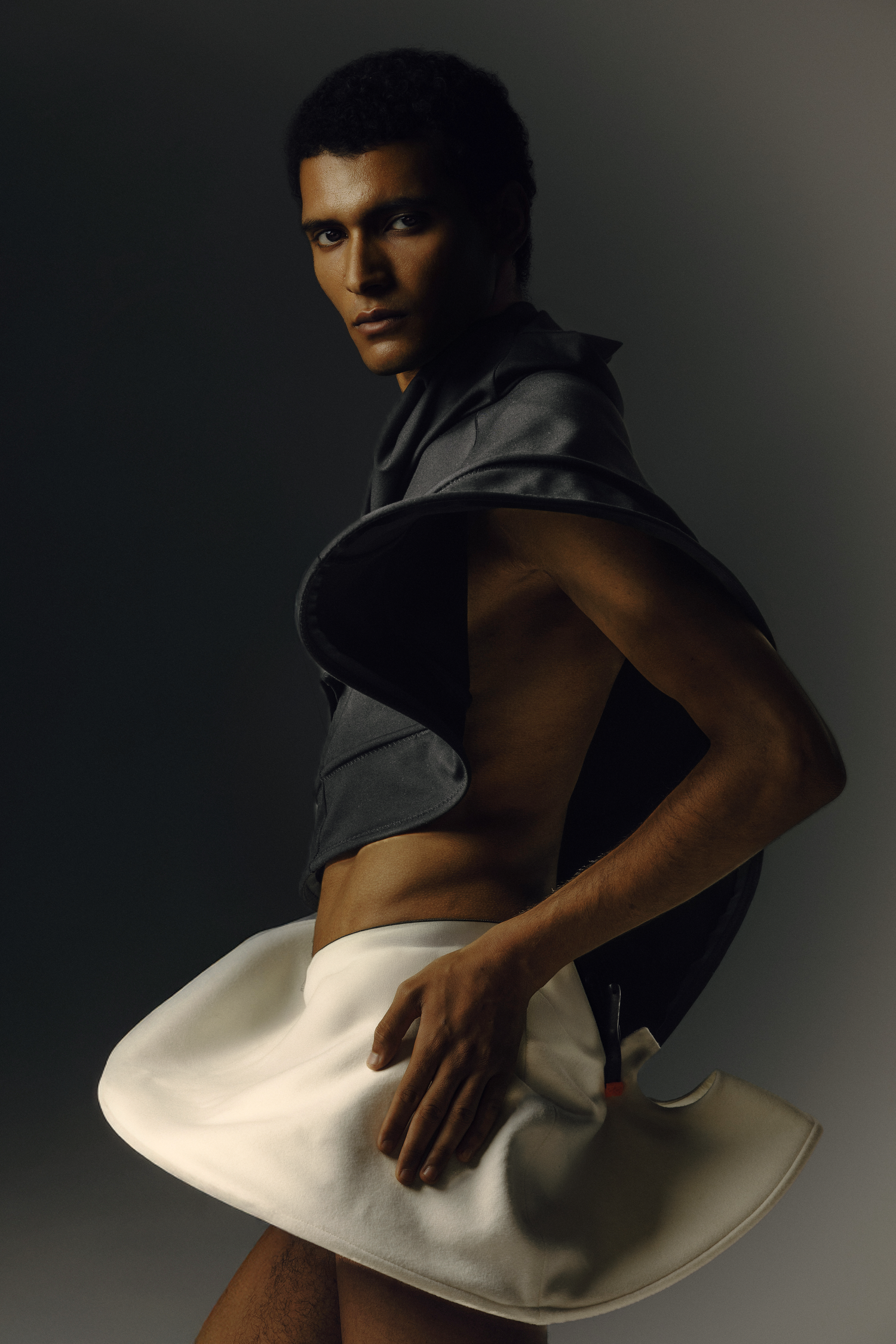 Male model wearing avant-garde structured gray jacket and white sculptural skirt in dramatic studio fashion shoot.
