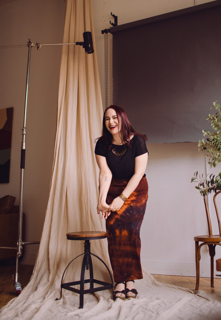 Woman in black tee and tie-dye maxi skirt laughs joyfully in photography studio with backdrop.