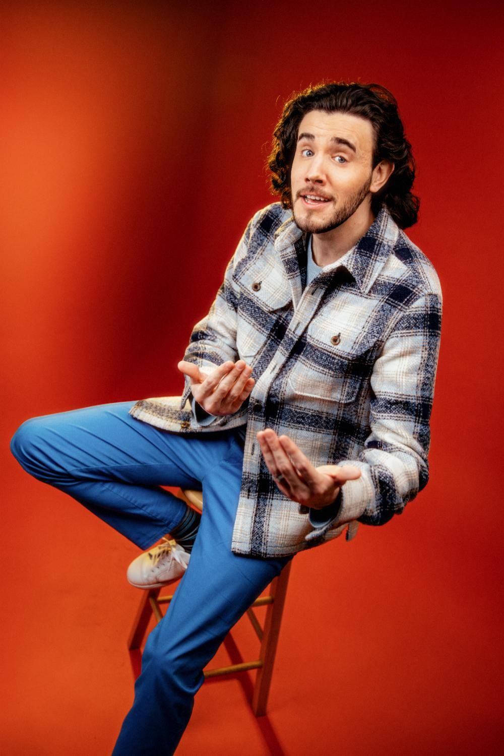 Curly-haired man in plaid jacket and blue pants sits on stool gesturing against red background.