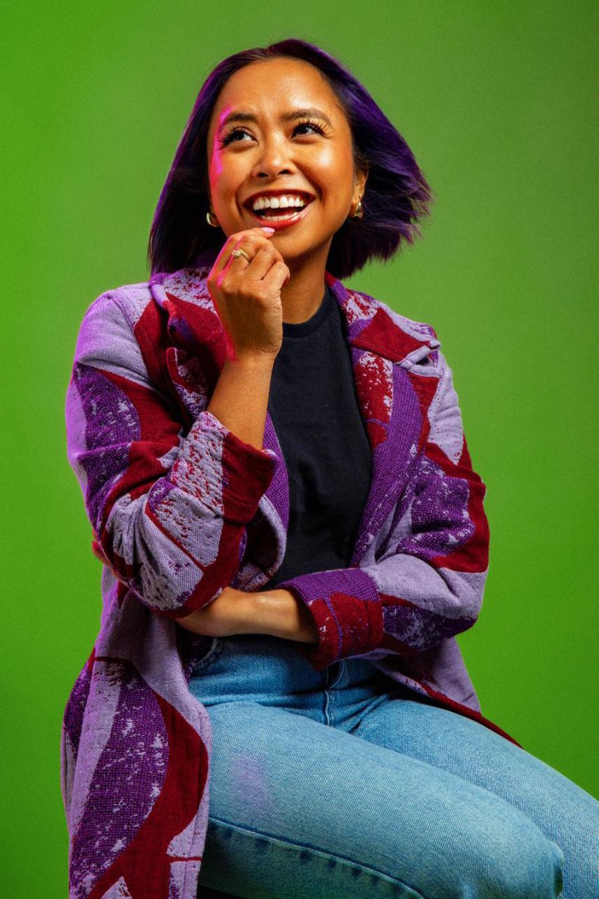 Laughing woman with purple hair wearing patterned purple jacket and jeans posing on green background.