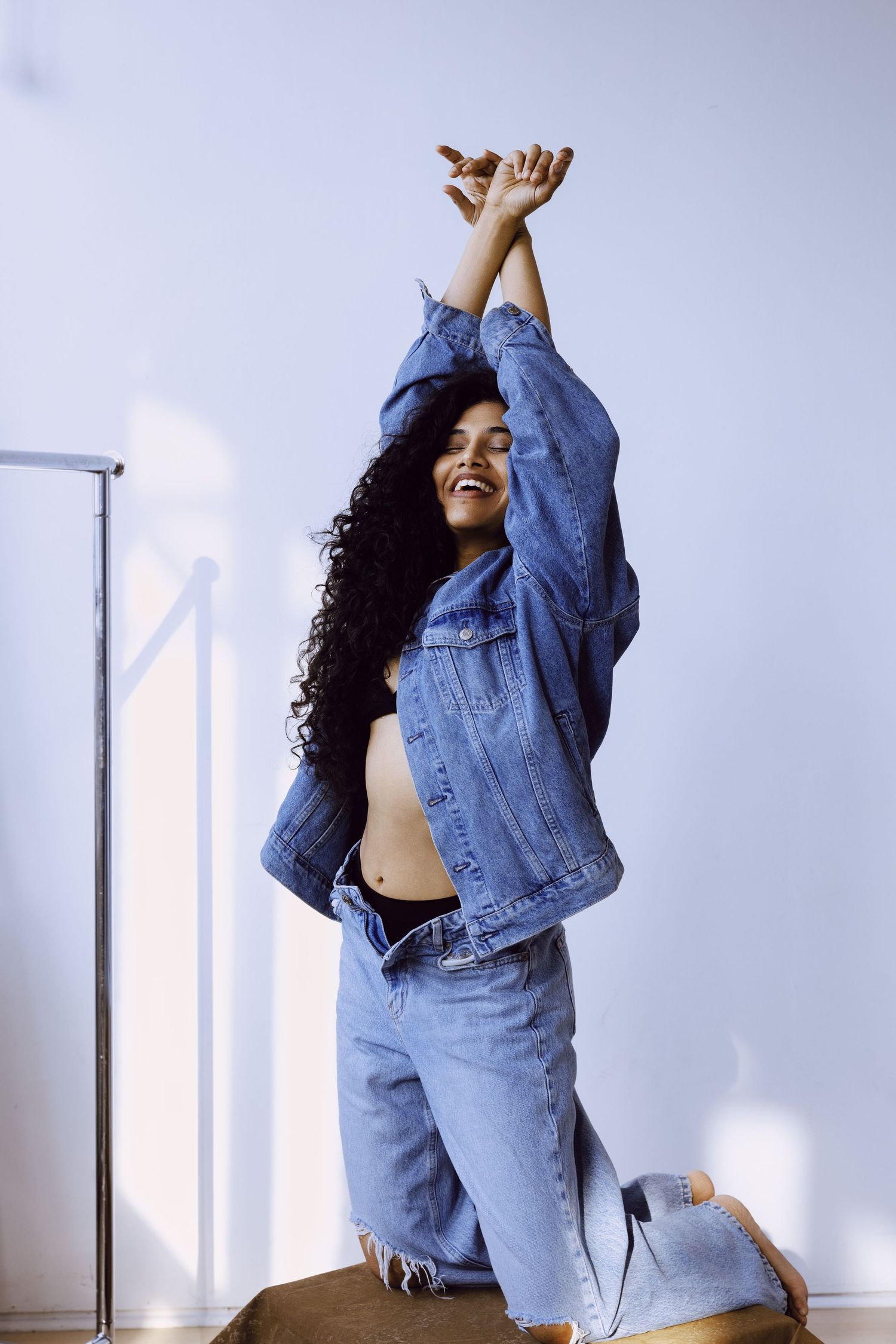 Young woman in denim jacket and jeans laughs with arms raised against light blue wall.