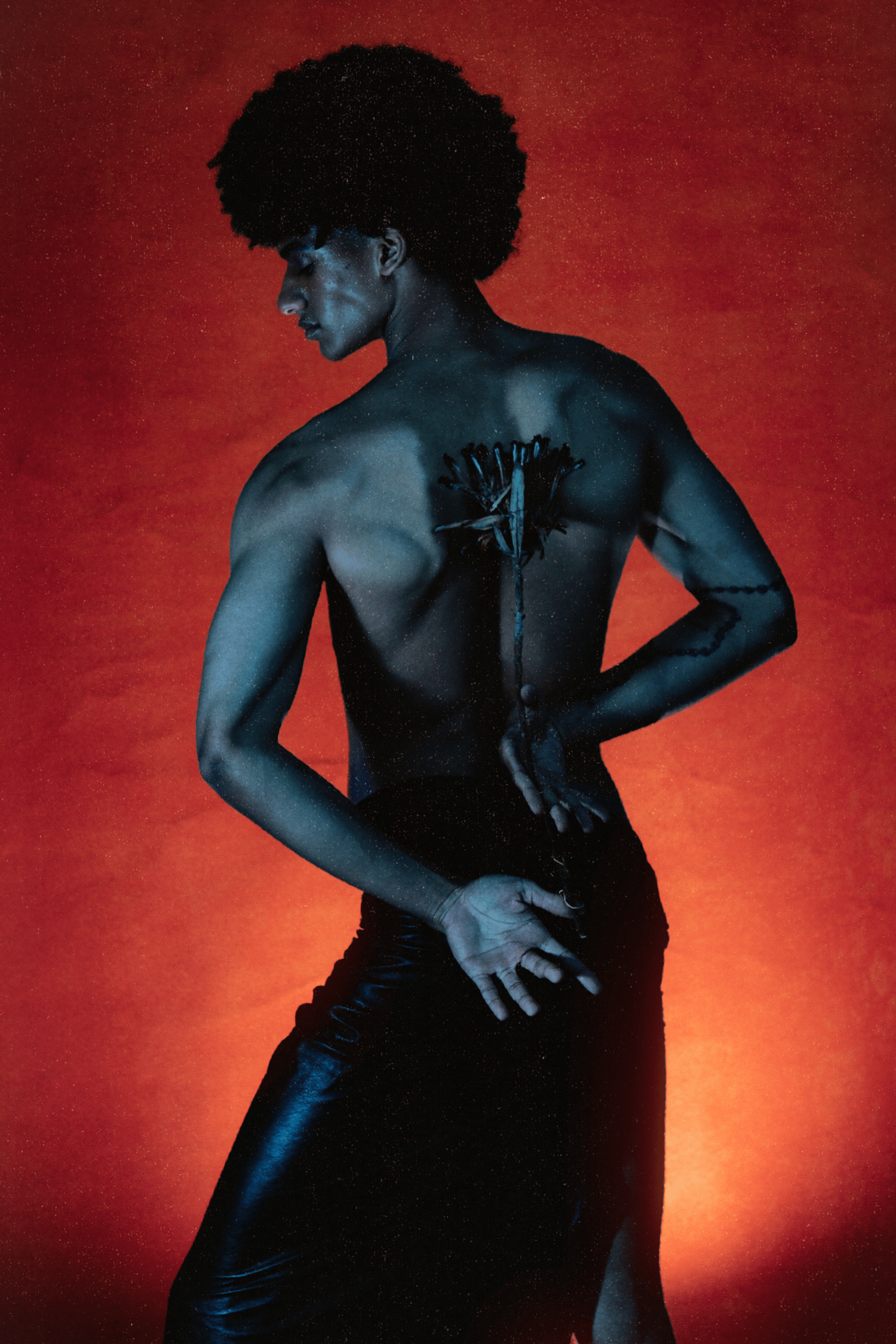 Black male model with natural afro in black leather skirt poses against dramatic red textured wall.