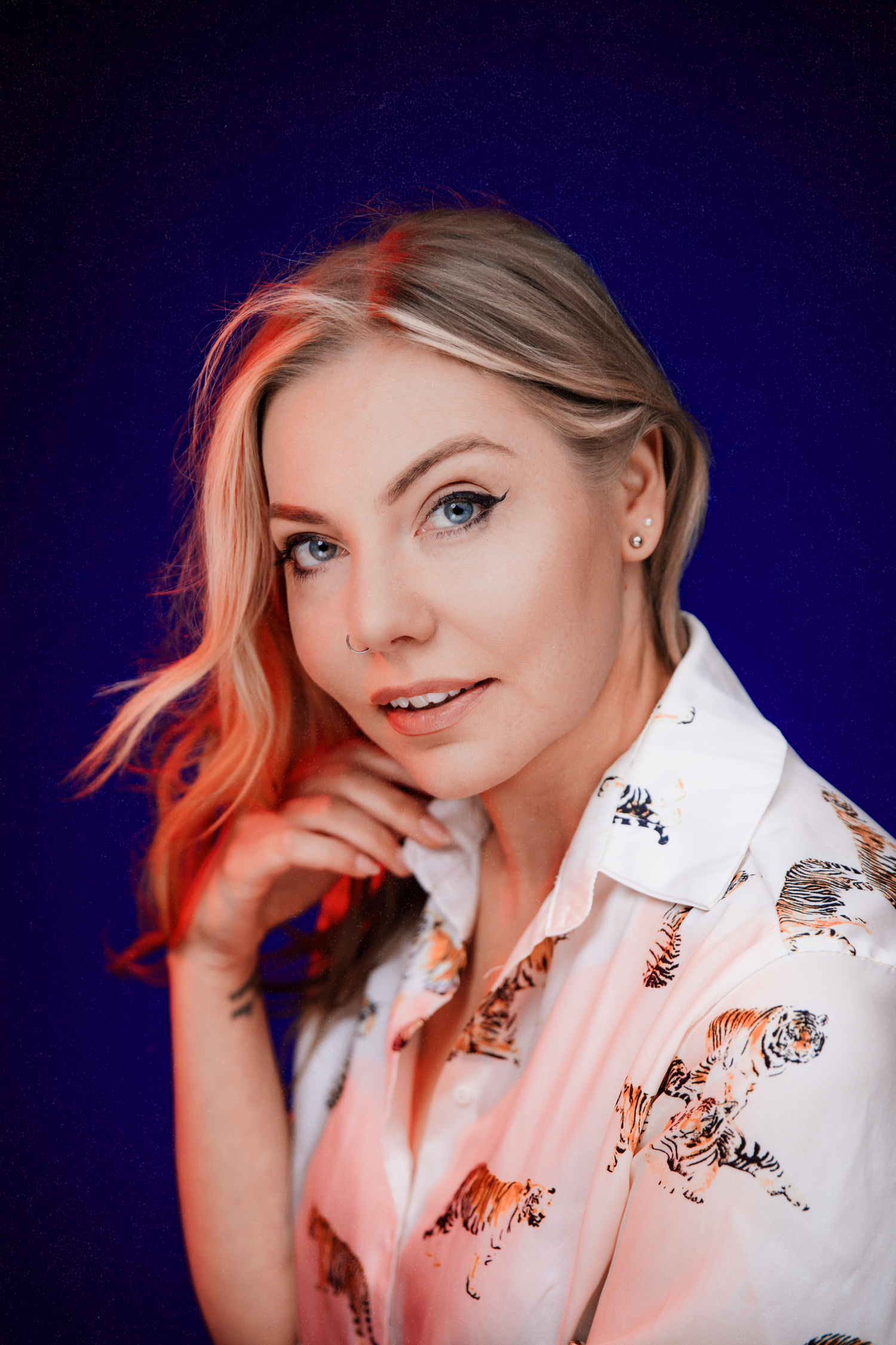 Young blonde woman in a white tiger-print blouse poses gracefully against a deep blue studio background.