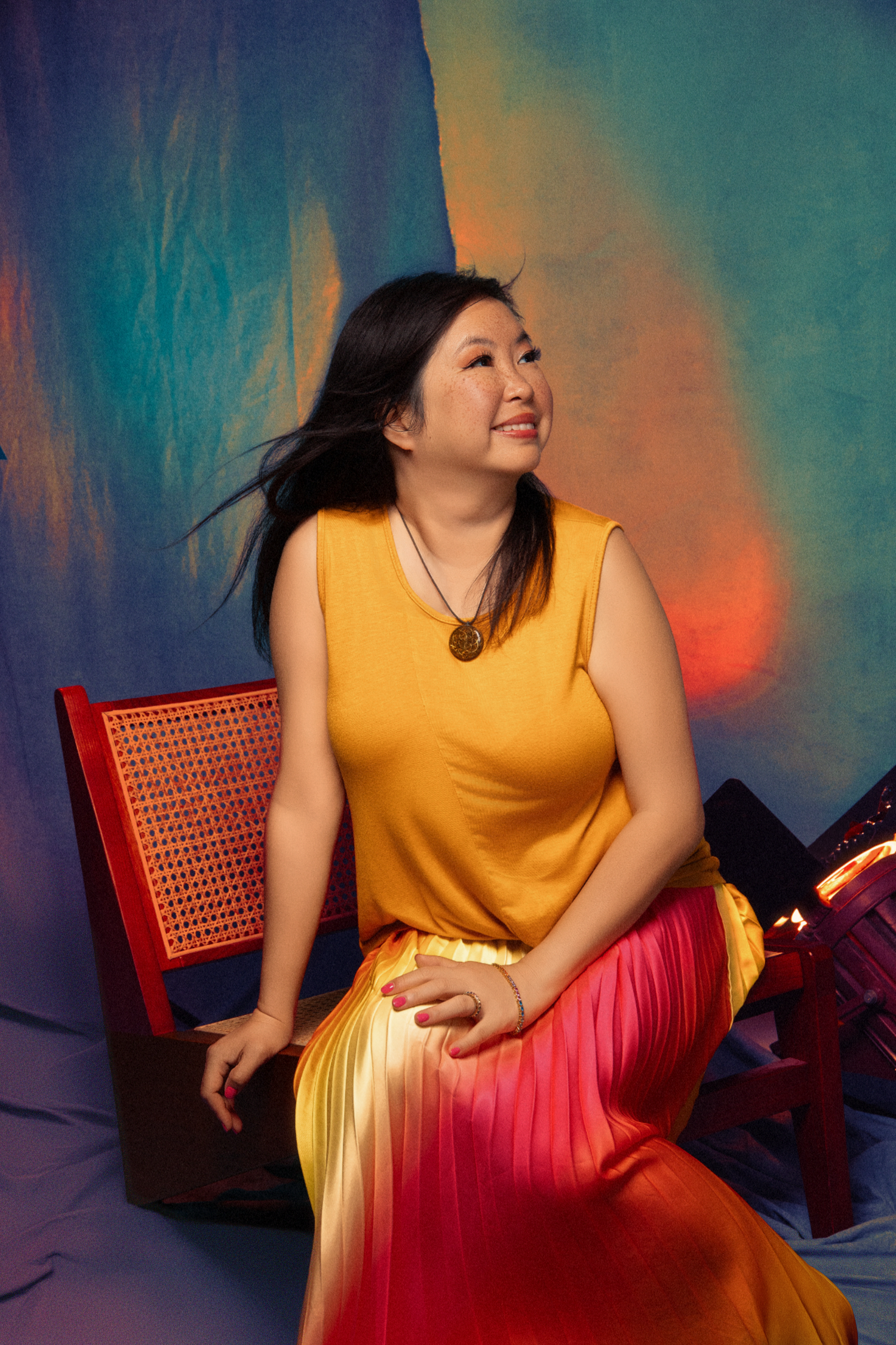 Woman in yellow top and gradient skirt sits smiling beside rattan chair against colorful backdrop.