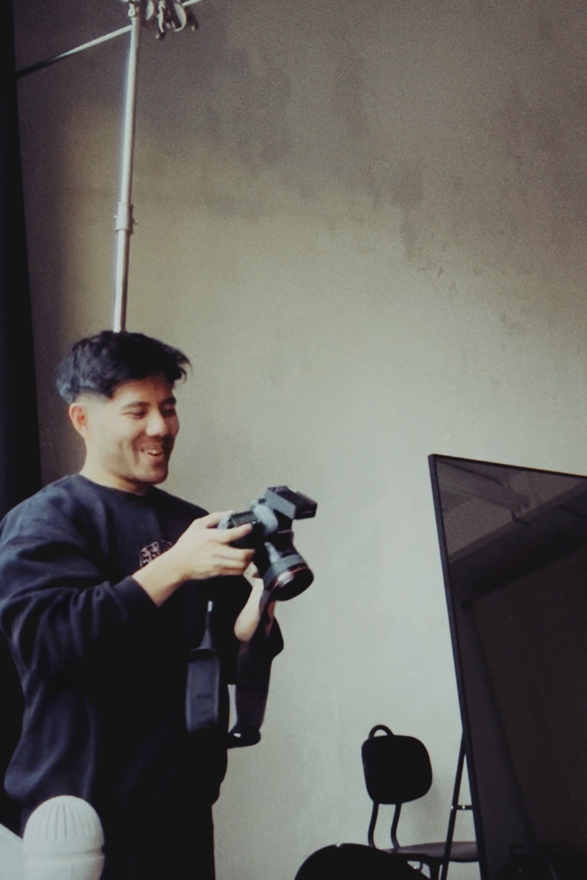 Smiling Asian male photographer reviews shots on camera in a photography studio setting.