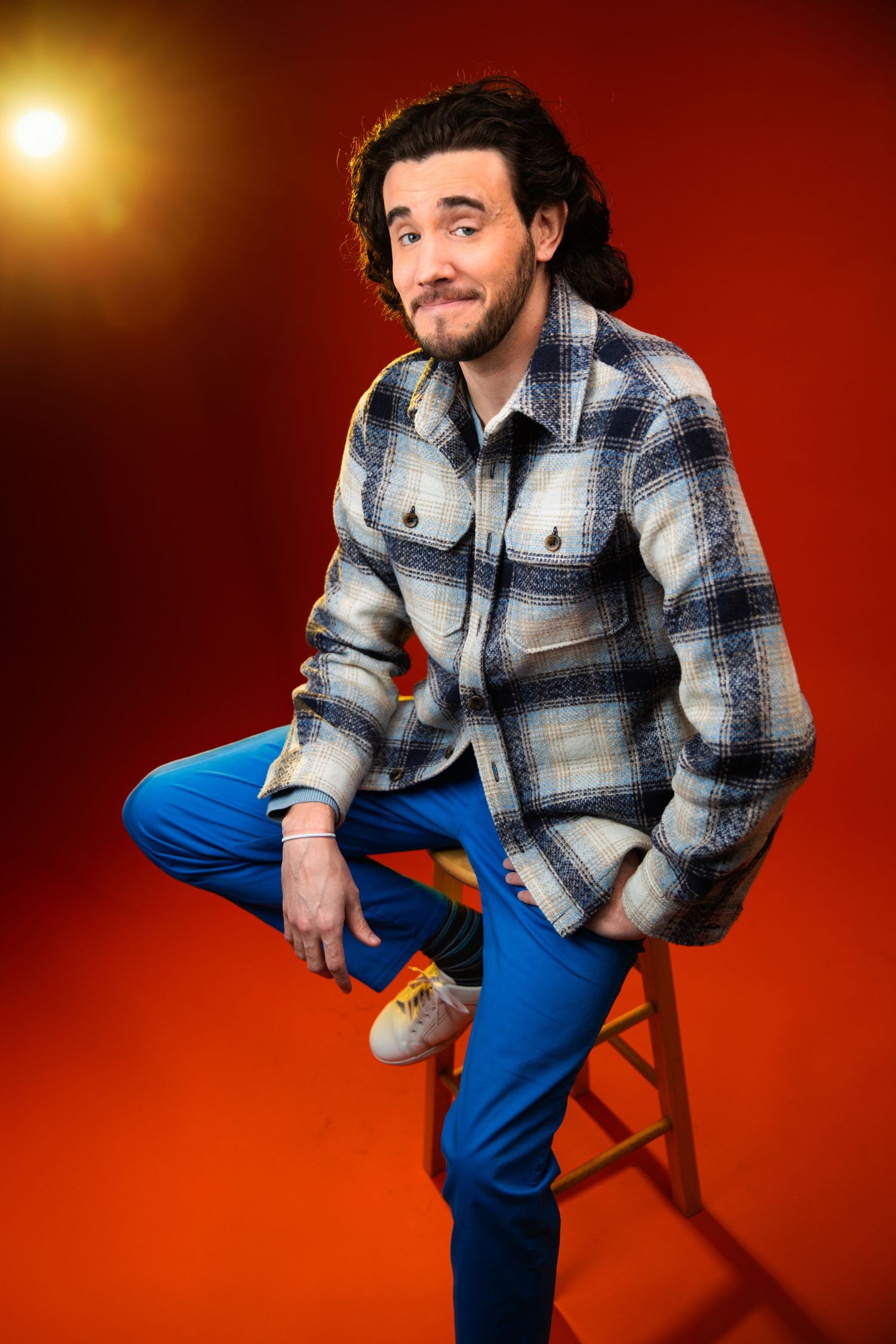 Stylish bearded man in plaid jacket and blue pants sitting on stool against bold red studio backdrop.
