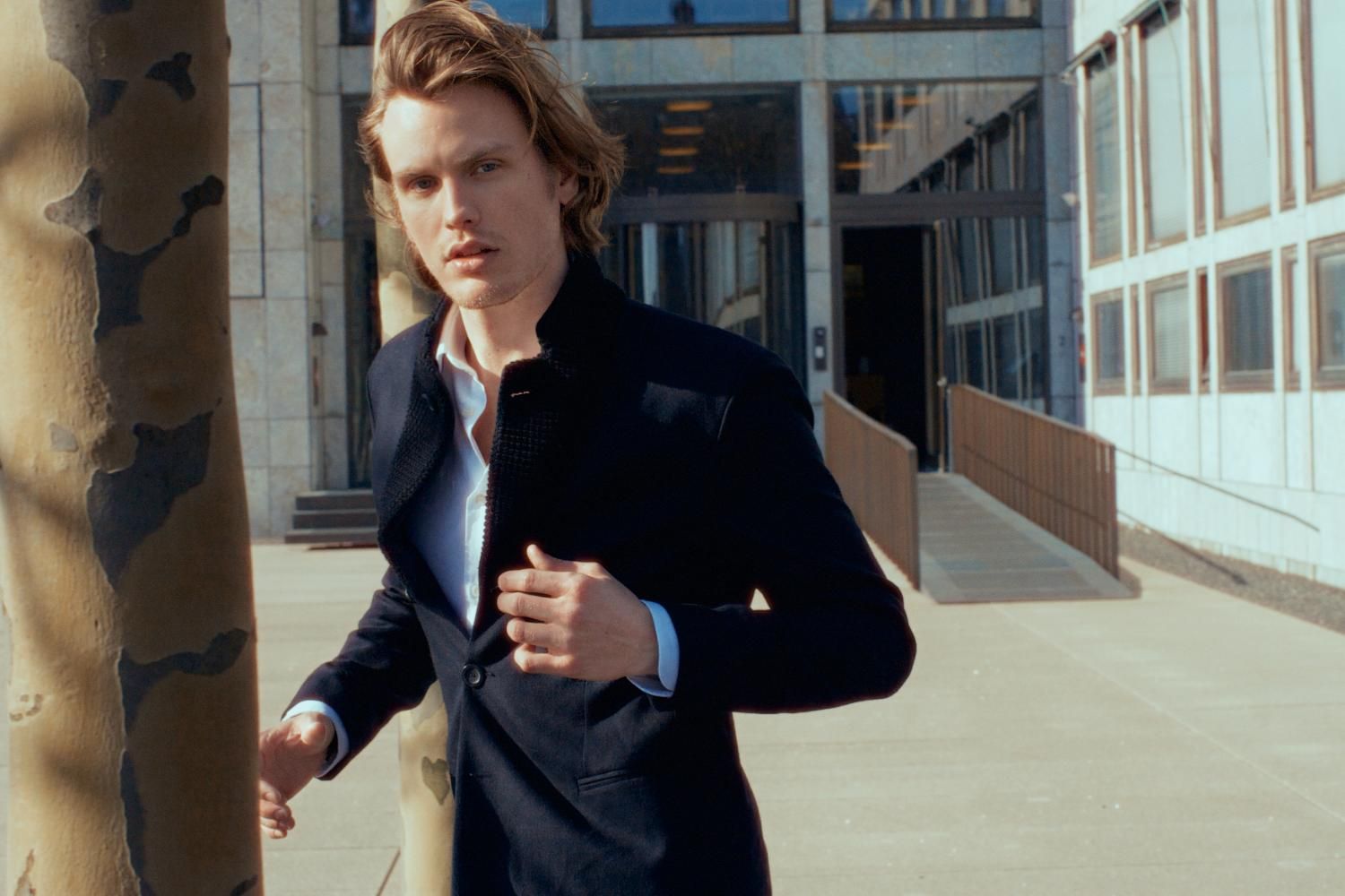 Young man in navy blazer and light blue shirt walks confidently past modern building exterior.