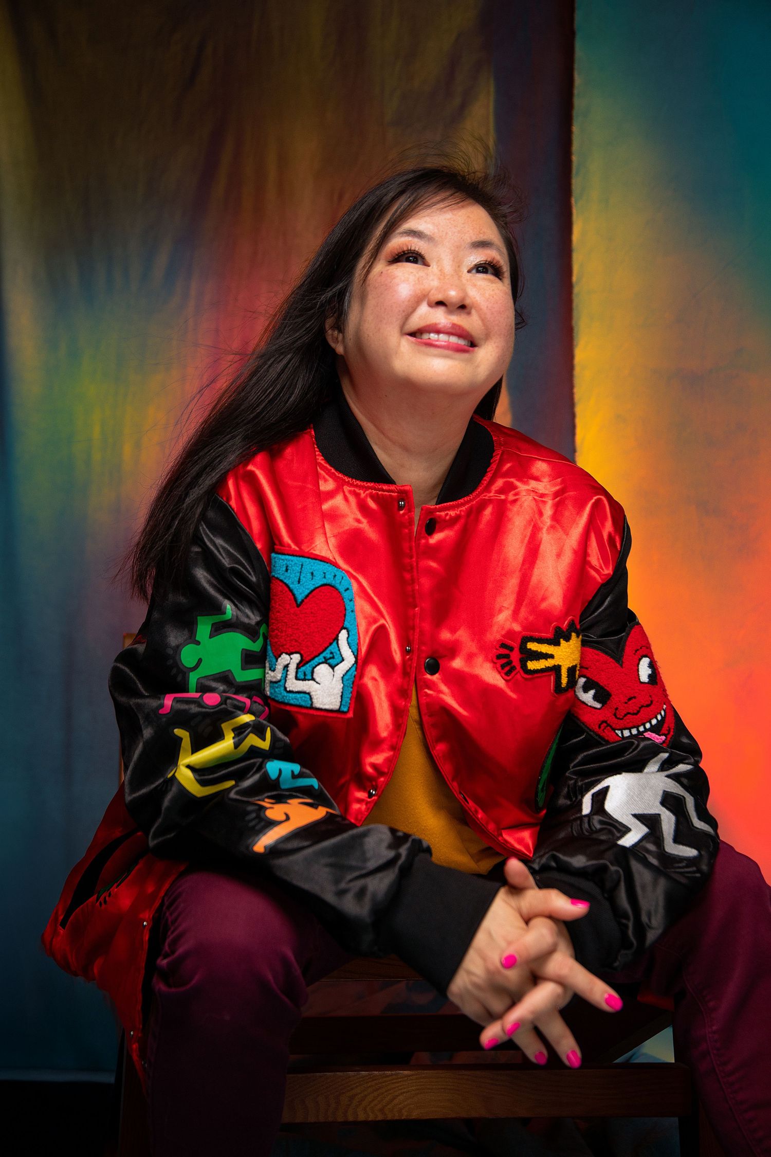 Woman in colorful Keith Haring bomber jacket sits smiling against vibrant painted backdrop.