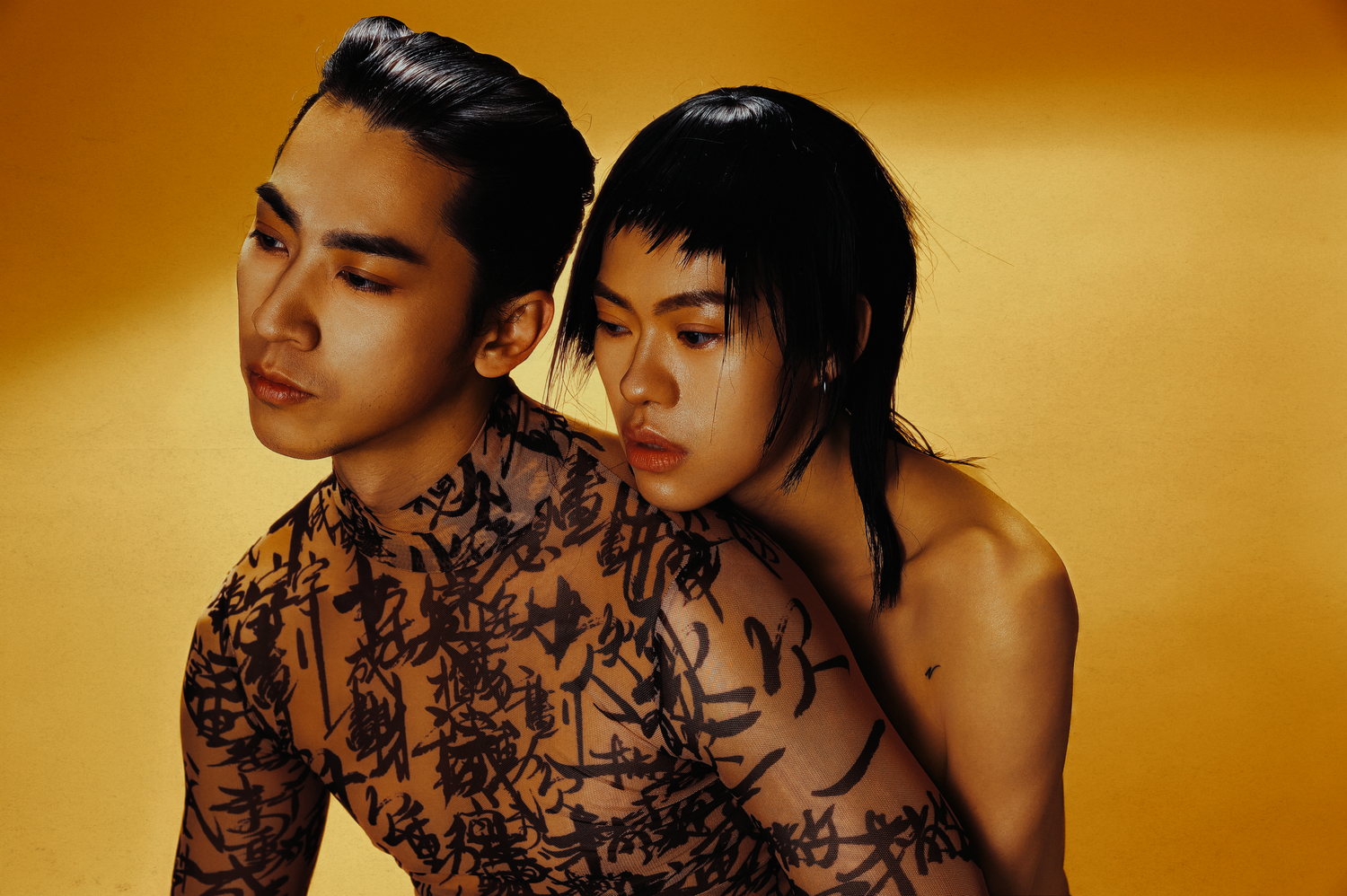 Two young people with artistic calligraphy body art pose together against a golden yellow background.