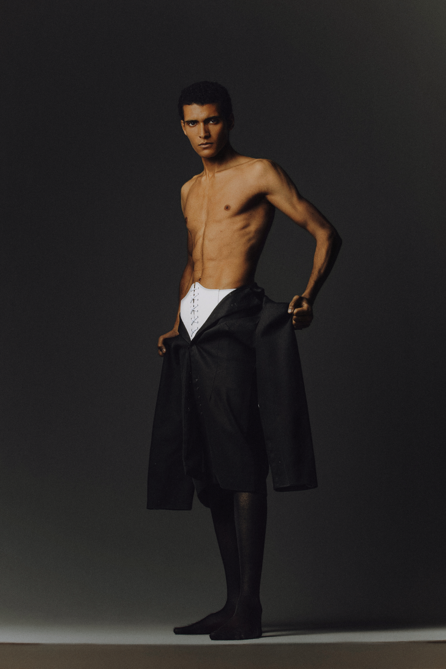 Shirtless male model in black skirt and white underwear posing confidently against a dark gray studio background.