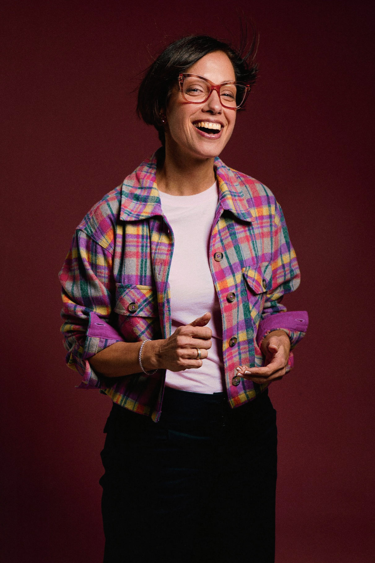 Laughing woman wearing red glasses and a vibrant multicolor plaid jacket poses against a dark red backdrop.