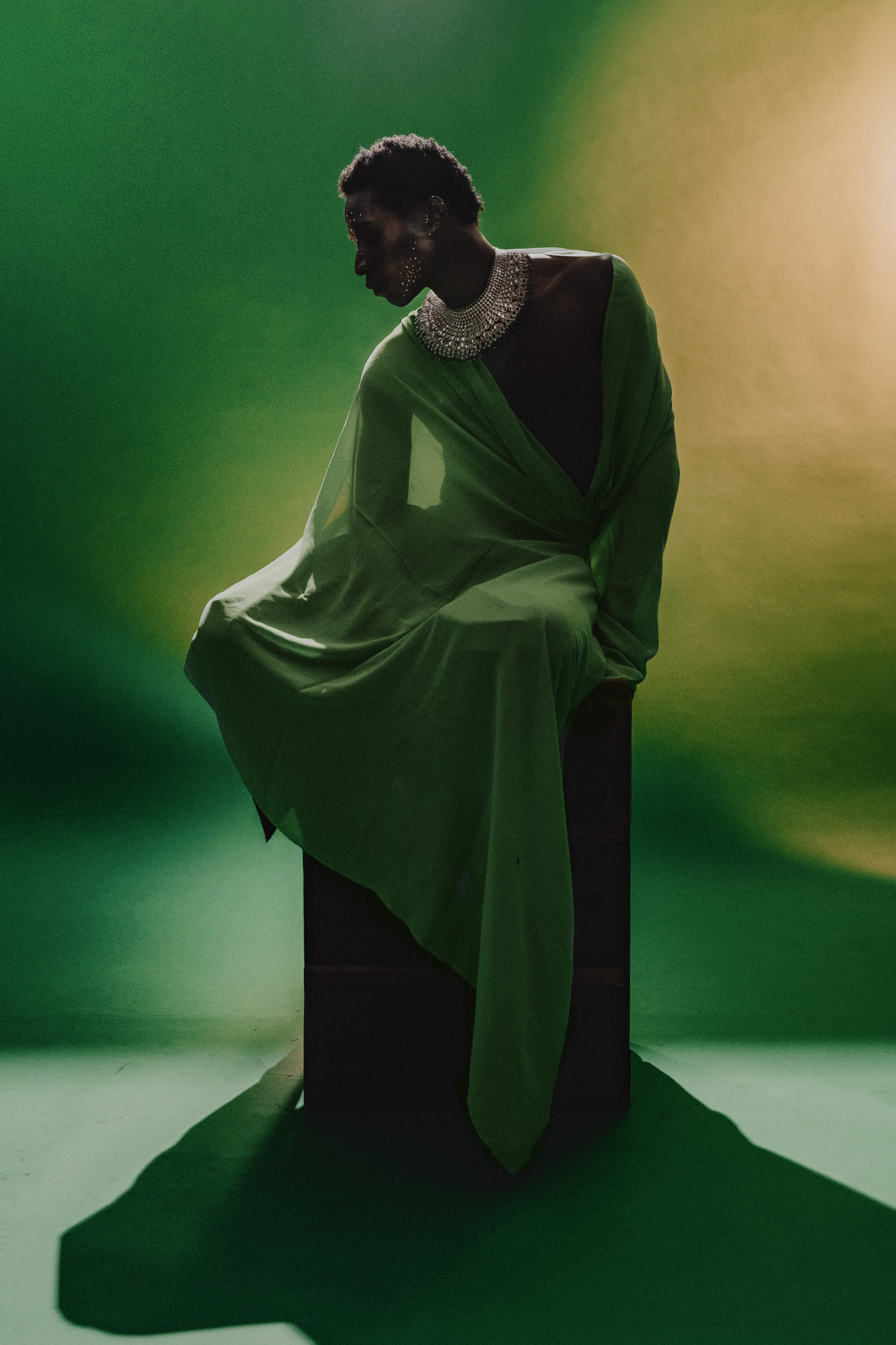 A Black model poses dramatically in a flowing green drape gown with pearl collar on a green backdrop.
