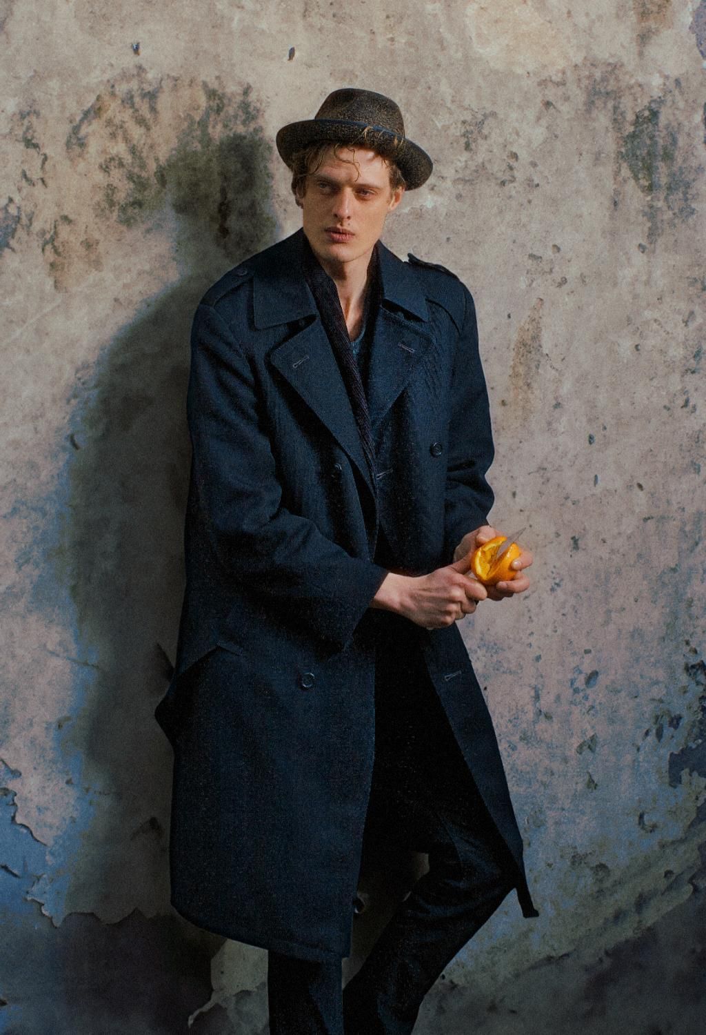 Young man in navy trench coat and fedora holding an orange against a textured wall.