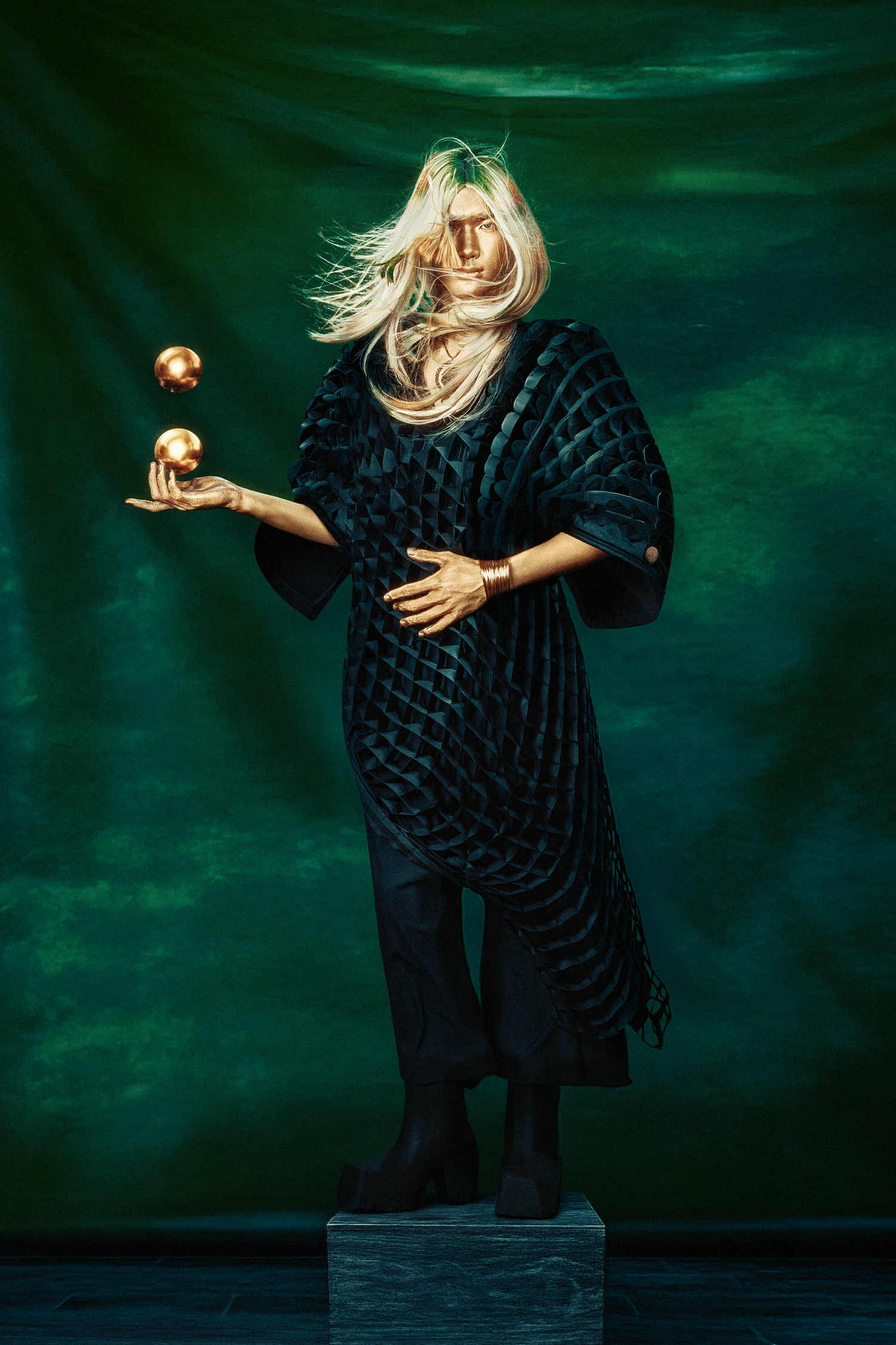 Blonde model in black velvet kimono holds golden orbs against emerald green backdrop.