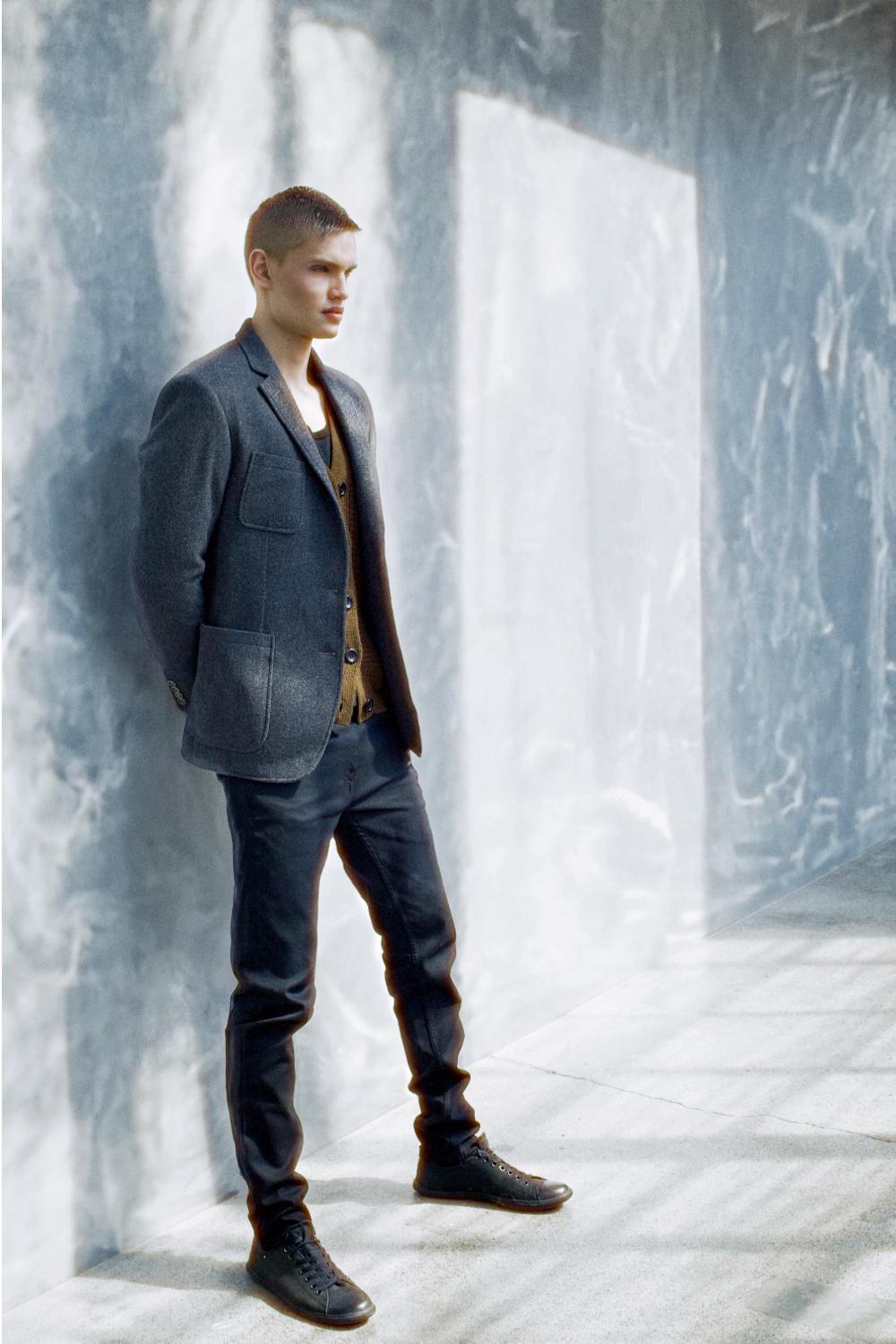 A young male model in a grey blazer and dark jeans poses in a softly lit marble-textured room.