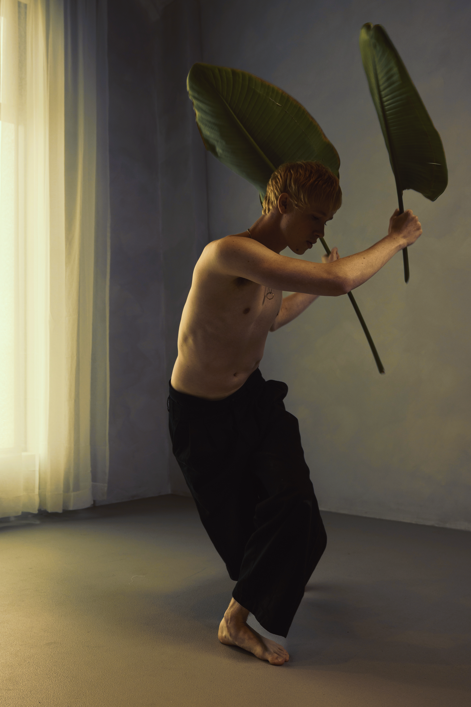 A shirtless man with blonde hair dances barefoot holding a large banana leaf in a softly lit studio space.