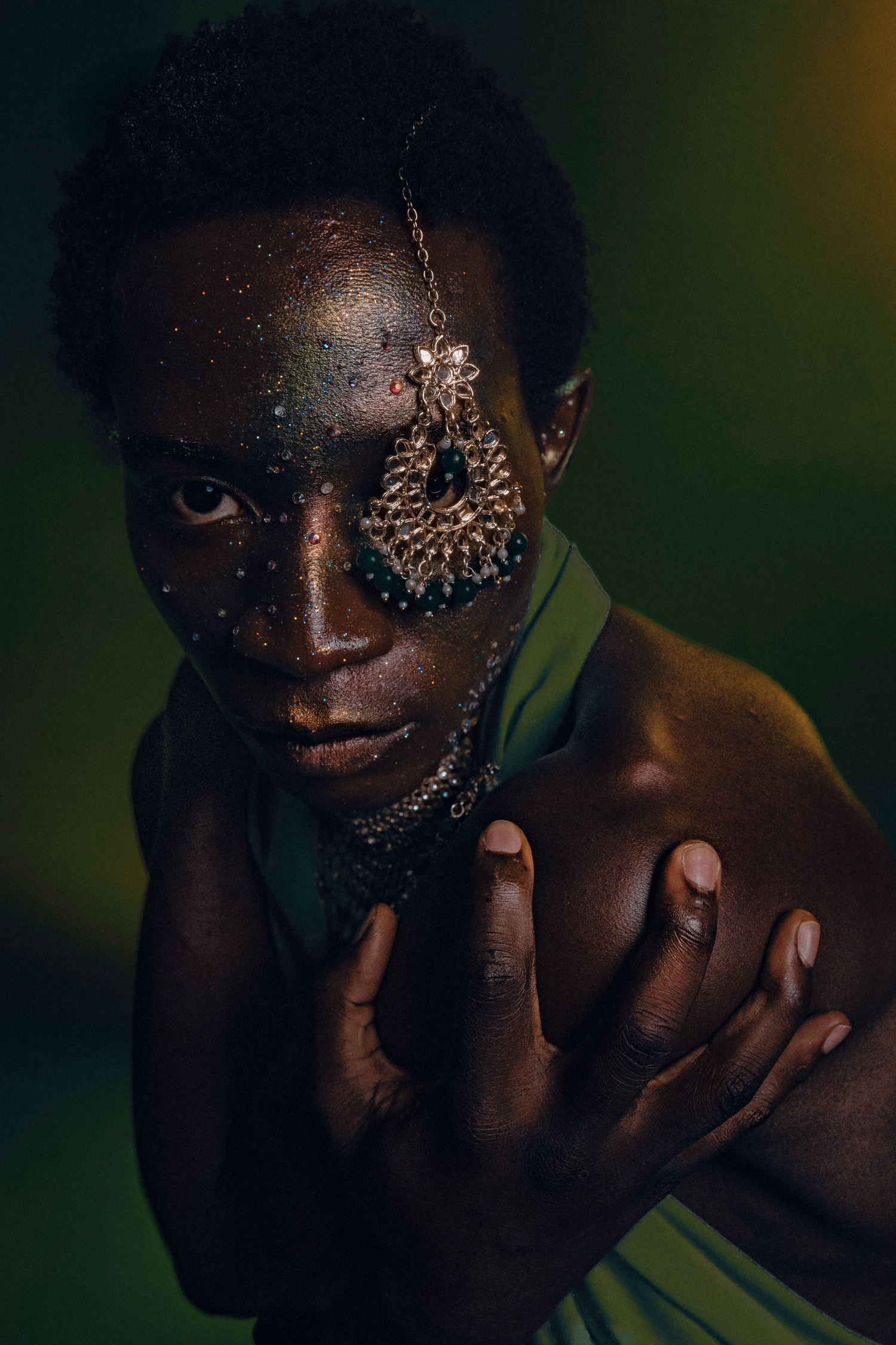 A Black model with glitter face makeup and ornate dangling earring poses dramatically against a dark green background.