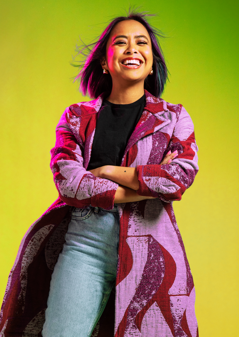 Smiling woman with multicolored hair in pink printed coat with arms crossed on yellow-green background.