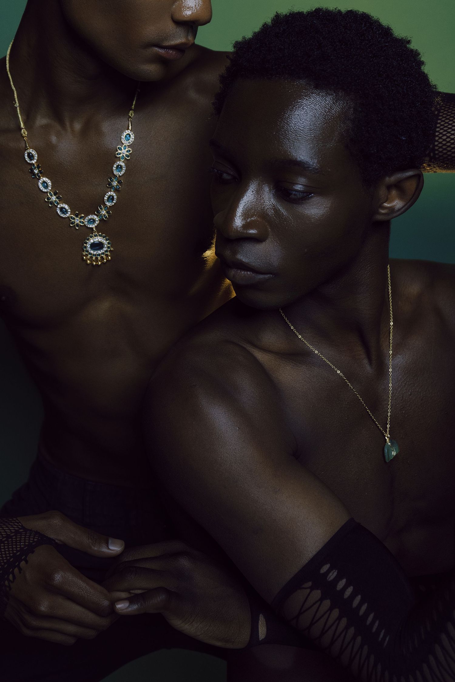 Two Black models in artistic jewelry shoot wearing gold necklaces against a moody dark green backdrop.