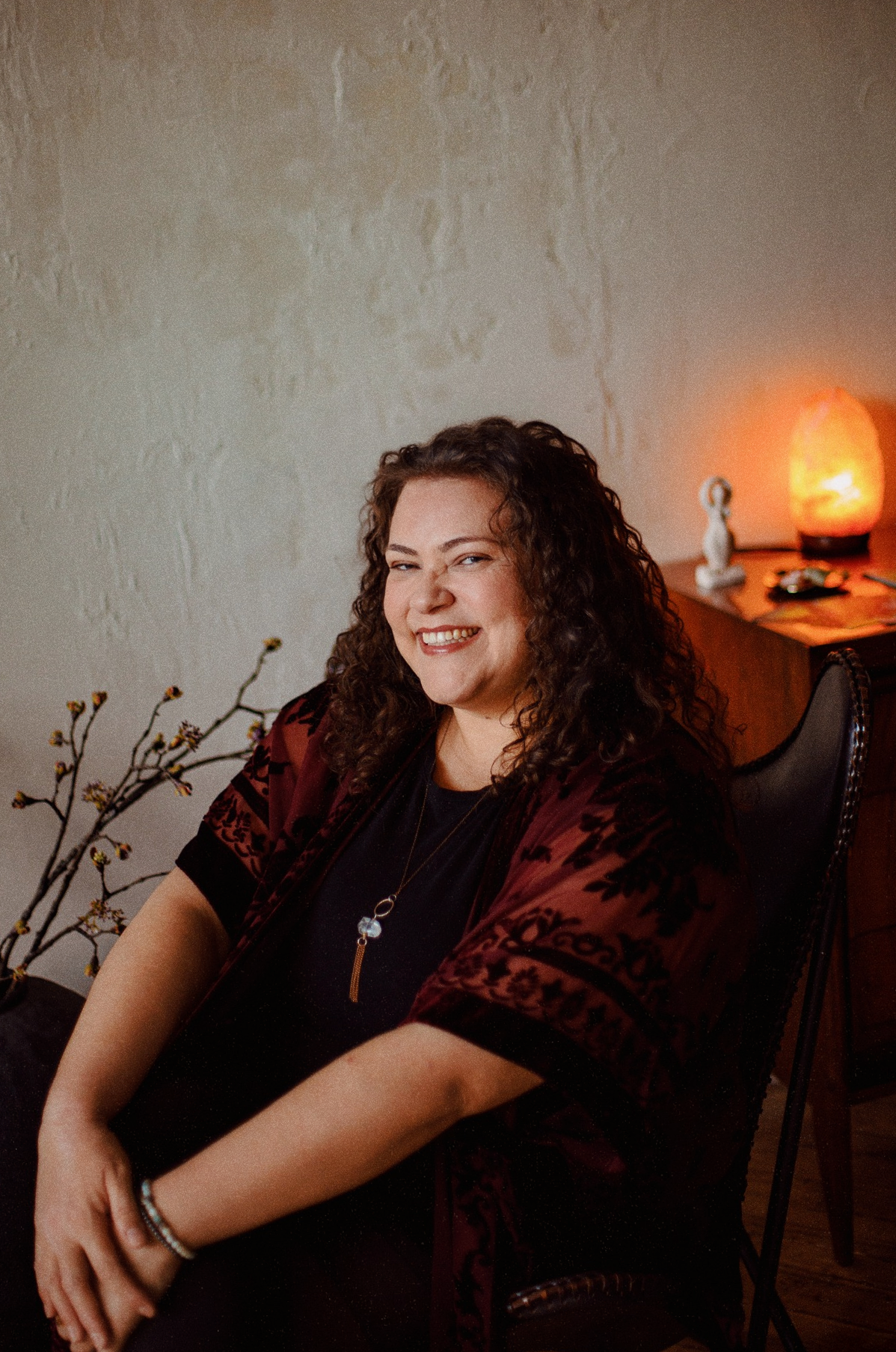 Curly-haired woman smiling warmly in a chair beside a glowing salt lamp.