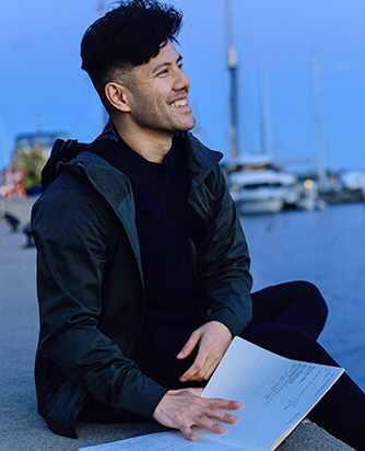 Young man smiling while sitting outdoors by marina at dusk holding sheet music papers.