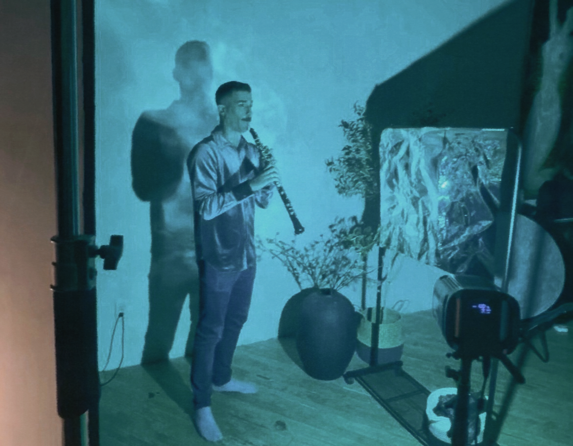 Male model holding clarinet stands in teal-lit photography studio surrounded by plants and equipment.