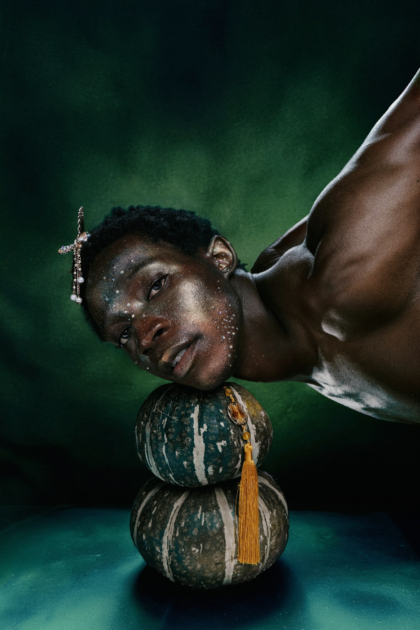 A shirtless Black man rests his head on stacked gourds, adorned with glitter and hair accessories, against a dark green background.