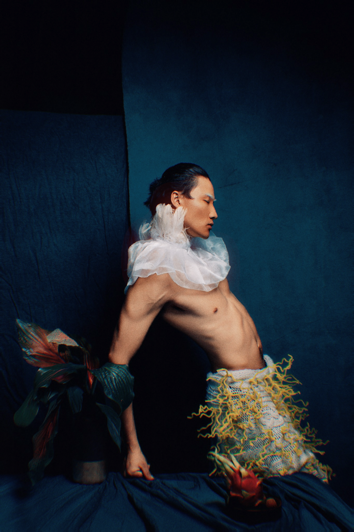 Shirtless Asian male model wearing white ruffled collar with colorful floral props on blue backdrop.