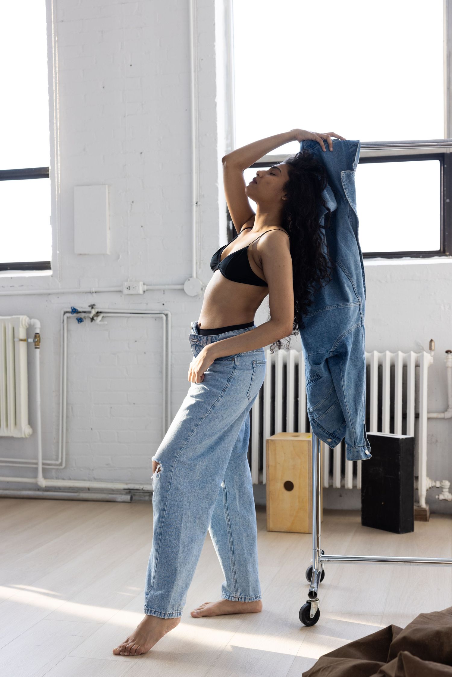 Woman in black bra and wide-leg jeans posing barefoot in bright studio with white brick walls and large windows.