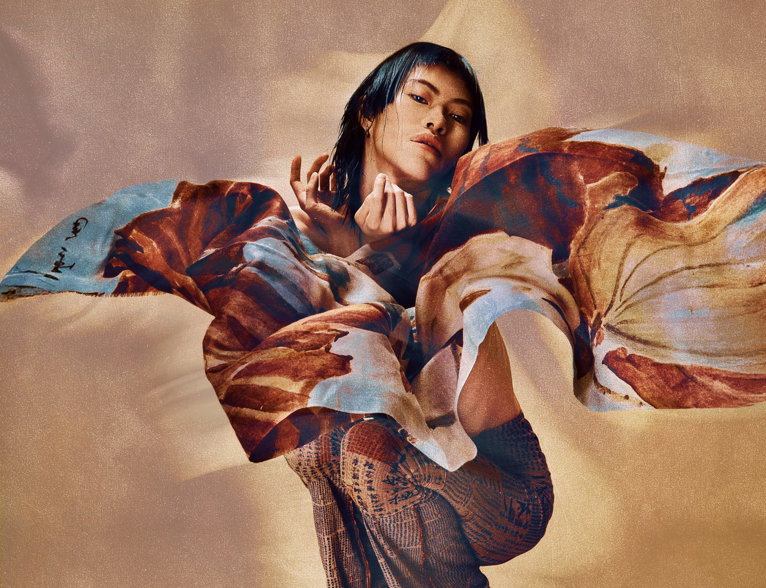 Woman in a flowing earth-toned abstract print outfit poses dynamically against a warm beige background.