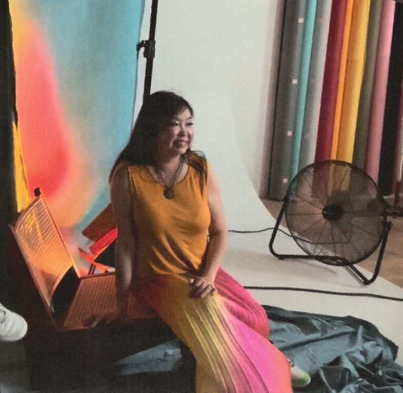 Woman in yellow dress sits smiling in colorful photography studio with fan and lighting equipment.