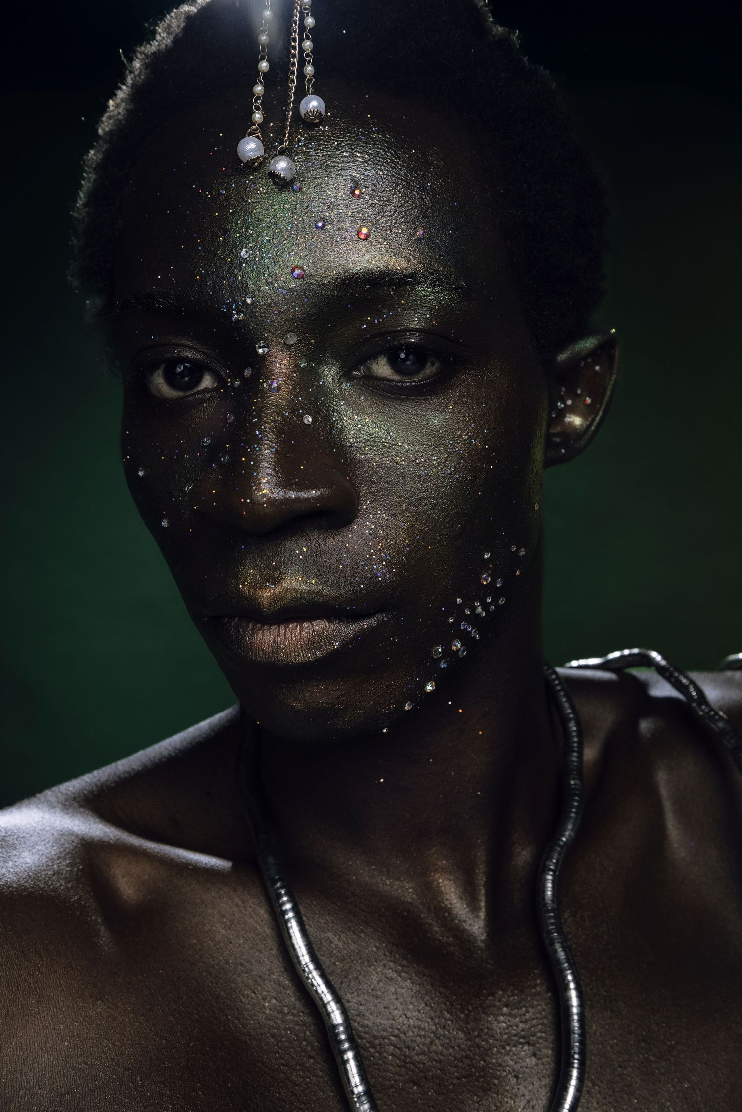 Black model with glitter face makeup and pearl head jewelry poses against a dark green background.