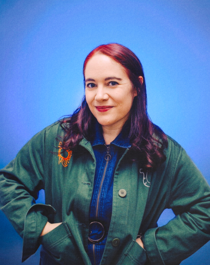 Woman with red hair in green jacket posing confidently against bright blue background with colorful earring.